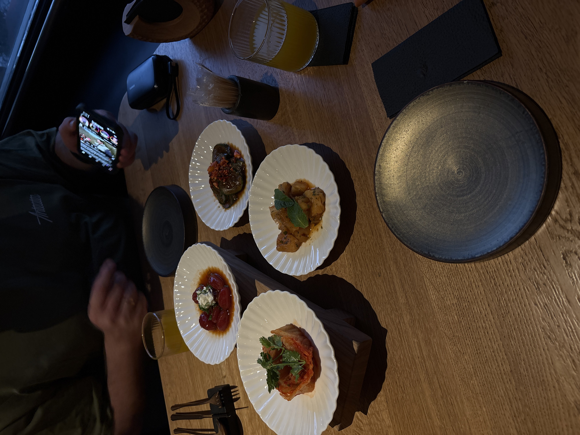 The image depicts a table laden with several small plates of food and a glass of juice, partially obscured by a person holding a smartphone, likely photographing the arrangement. The table is made of wood and displays four plates with distinctive scalloped edges. The plates contain various dishes; one with what appears to be tomato and cheese, another with braised cabbage topped with fresh cilantro, a third with sautéed or roasted potatoes and fresh mint, and a fourth with meat and vegetables. Utensils are placed next to the lower left plate.
Next to the dishes are a black container with what seem to be straws or toothpicks, a glass of orange juice, and a dark grey plate of a similar size to the others. A black square cloth sits next to it.
The person holding the phone is wearing a dark green shirt, and the phone's screen is visible, displaying what seems to be an Instagram feed. The camera and additional black accessories can be seen to the upper-left corner of the image.
Based on the menu and interior design style, this could be a trendy restaurant. Given the stated location (Krasnoyarsk, Russia), it is reasonable to assume the restaurant is in this city.