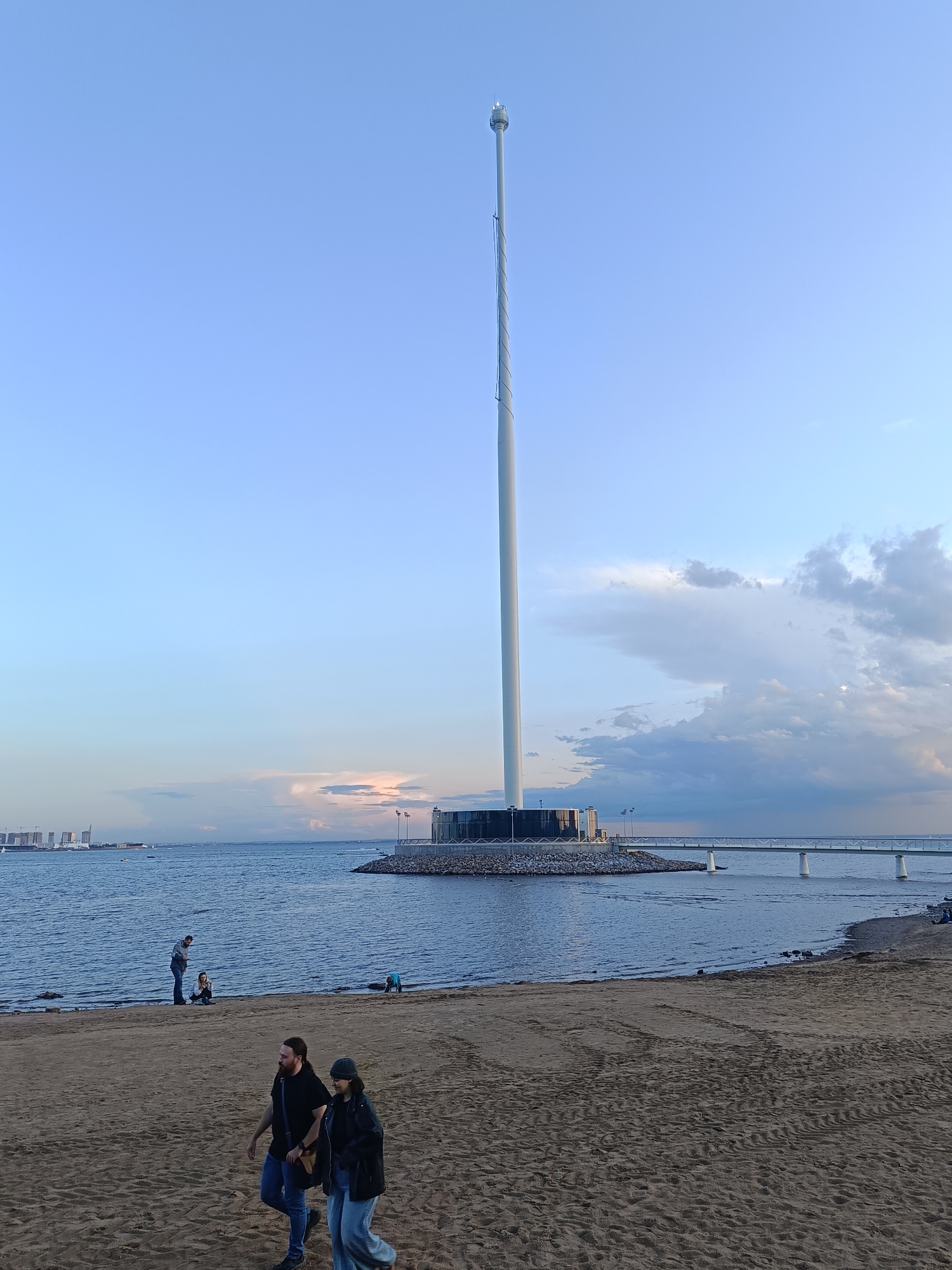 The image depicts a wide shot of a beach scene with a tall, slender tower dominating the background. The tower, a prominent structure, is white and tapers to a point, topped with what appears to be a beacon or antenna. It stands on a circular platform surrounded by water. To the right of the tower, a modern building with dark glass windows is visible, connected to the shore by a bridge. The sea is calm, with a few small boats dotting the surface. The sky is a pale blue with scattered clouds, some of which are tinged with sunset colors.
In the foreground, a sandy beach stretches across the bottom of the frame. Several people are present on the beach and by the water's edge. Two individuals are walking towards the camera on the sand, a man with a beard and dark clothing, and a woman in a dark jacket and beanie. Further back, by the water, a group of three people are gathered, with one person standing and two sitting. Another individual in a blue top is crouched near the water. The beach shows tire tracks, suggesting recent activity from a vehicle. In the far distance, a cluster of tall buildings can be seen on the horizon.
This scene is the Kurchatov Institute's meteorological tower, located in the Primorsky District of Saint Petersburg, Russia. The tower is part of a complex near the Gulf of Finland.