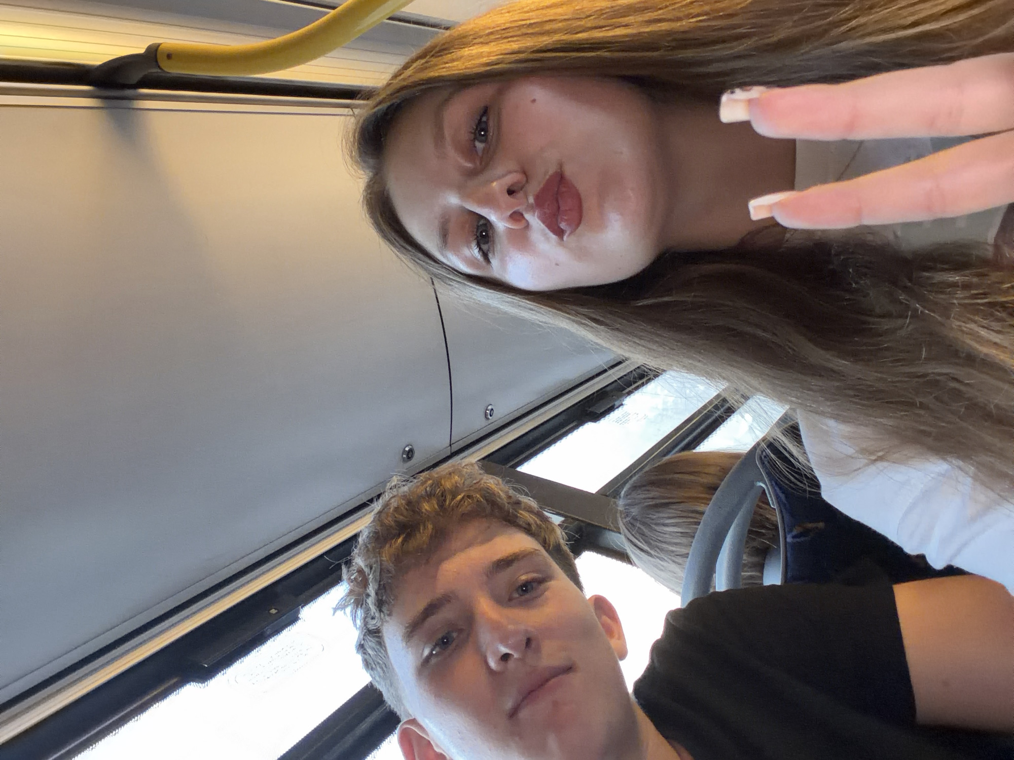 A young Caucasian man with short, curly blonde hair is in the foreground on the left, looking slightly down and to the left. He is wearing a black t-shirt. Next to him on the right, a young Caucasian woman with long, light brown hair is making a duck face and holding up two fingers in a peace sign. She is wearing a white top. Behind them, another person's head with blonde hair is visible. The scene appears to be inside a public transportation vehicle, possibly a bus, with a yellow handrail visible in the upper right corner and windows on the left side. The interior of the vehicle has a light-colored ceiling and side panels. The lighting suggests it is daytime, possibly overcast. There is no indication of the location being Burgas, Bulgaria.