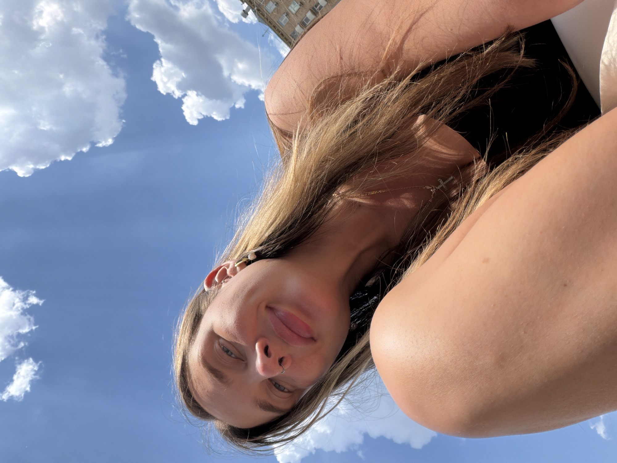 The image shows a close-up, low-angle selfie of a young woman outdoors with a cloudy blue sky in the background, and the corner of a building visible at the top. The woman has long blonde hair, a nose ring, and is wearing jewelry, including a cross necklace and earrings. She is looking towards the camera with a slight smile. The lighting suggests it is daytime, likely sunny with scattered clouds. The setting appears to be an urban environment, given the building in the background. The woman's relaxed posture and smiling expression convey a sense of leisure and happiness. The image is oriented sideways.