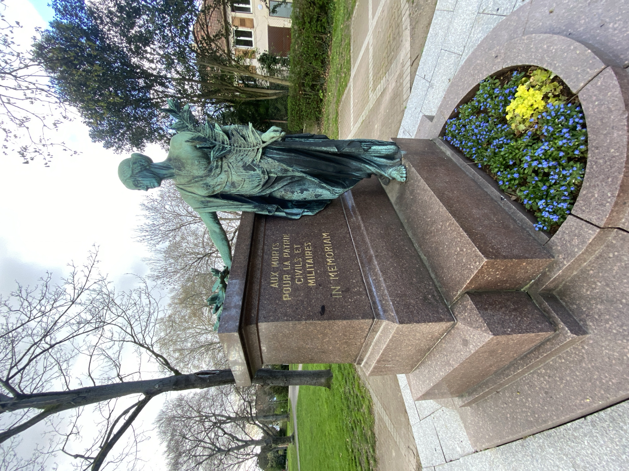The image shows a war memorial in Schiltigheim, France. The monument consists of a bronze sculpture of a female figure holding a laurel branch, placed on a multi-tiered stone base. The inscription on the base reads "AUX MORTS POUR LA PATRIE CIVILS ET MILITAIRES IN MEMORIAM". A flowerbed with blue and yellow flowers is situated next to the statue. The memorial is located in a park setting, with trees, grass, and a paved pathway visible in the background. Buildings are visible in the distance. The image is taken on a partly cloudy day.