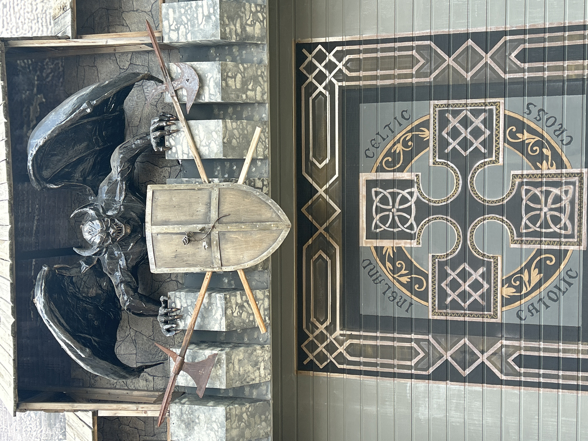 The image shows a facade of a building with a gargoyle-like sculpture perched above a large, painted Celtic cross design on a garage door. The sculpture, made of dark metal, depicts a creature with bat-like wings, sharp teeth, and clawed hands, gripping a wooden shield. Two rusty battle axes are positioned on either side of the shield, leaning against the faux stone structure behind it. The lower part of the building features a gray, corrugated garage door adorned with a large, intricate Celtic cross in shades of gold, black, and light gray. The words "IRELAND," "CELTIC," and "CROSS" are visible, encircling the cross design. The architectural style of the upper section suggests a medieval or fantasy theme, possibly a themed establishment. This structure is located on Maly Pestrinskoy Street in the Sverdlovsk District of the city of Perm, Russia.