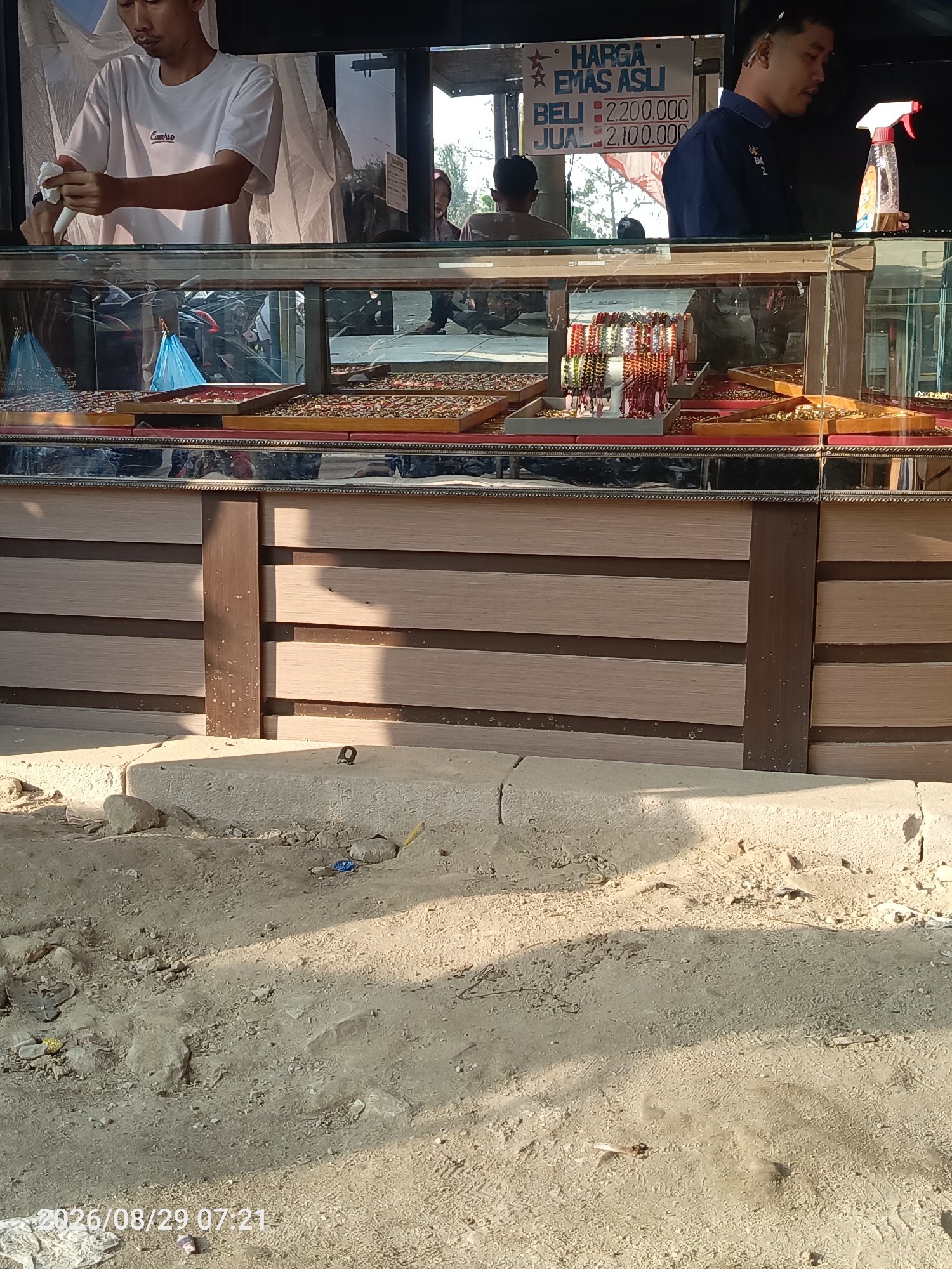 The image depicts a street-level view of a small jewelry stall in Sangiang, Indonesia. In the foreground, a rough, sandy ground is visible, scattered with rocks and debris, with a concrete ledge forming the base of the stall. The stall itself features a wooden structure with horizontal planks, topped by a glass display case filled with various gold jewelry. Among the displayed items are rings and bracelets. Behind the glass, two men are visible. One man, wearing a white t-shirt, stands on the left, appearing to be cleaning or preparing something. The second man, wearing a dark blue shirt, is on the right, standing near a spray bottle and a sign that reads "HARGA EMAS ASLI BELI: 2.200.000 JUAL: 2.100.000". This sign indicates the prices for buying and selling authentic gold. The background suggests an outdoor environment with some foliage and a bright sky.
