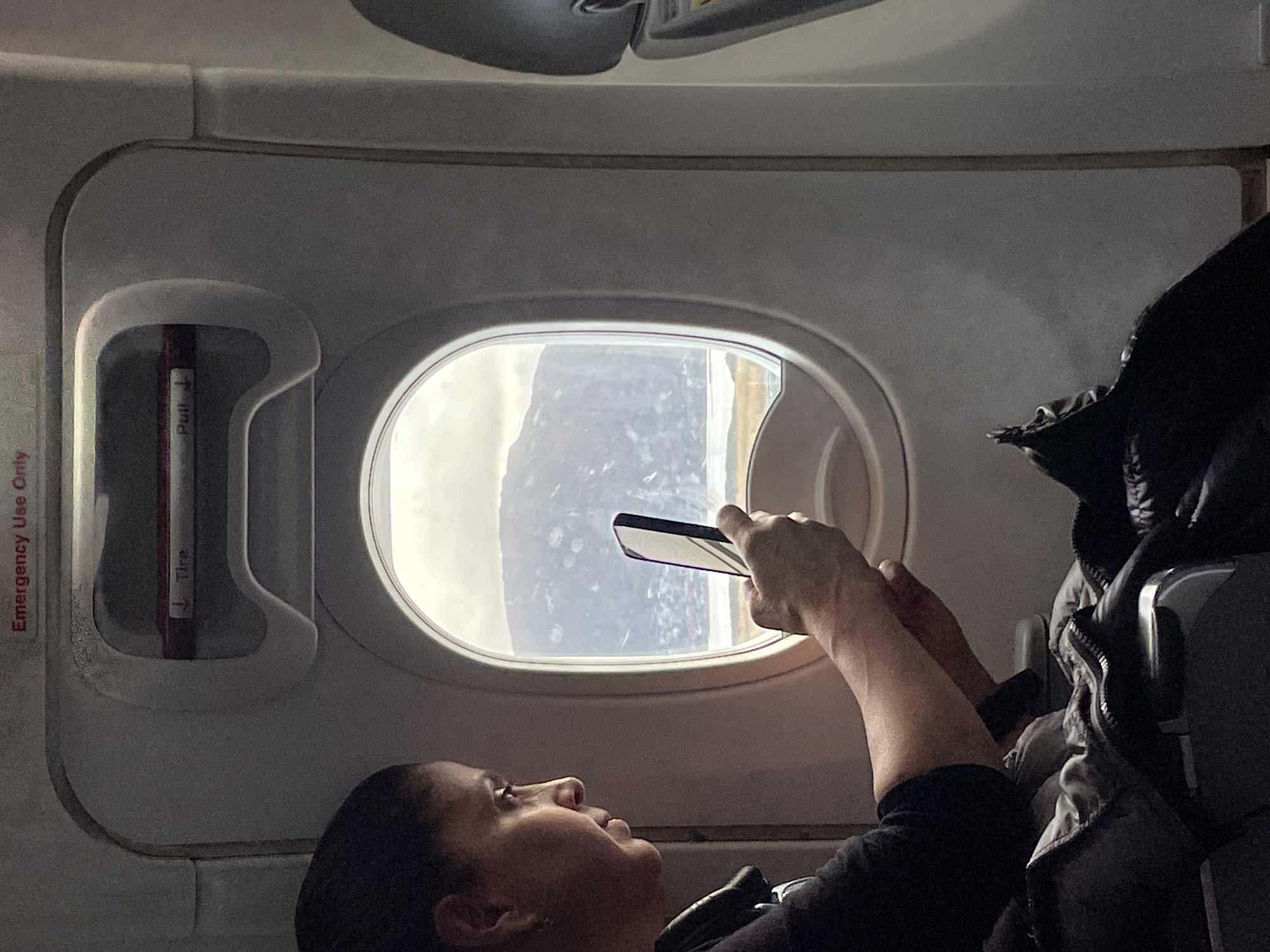 An indoor, eye-level shot captures a person inside an airplane cabin, attentively focusing on the view outside a window. The subject, a person with dark hair, is seen in profile from the chest up, positioned towards the left of the frame. They are holding a black smartphone with both hands, extending it towards the elliptical airplane window, appearing to take a photograph or video of the scenery beyond.
Through the window, a bright, possibly overcast sky is visible, with indistinct shapes of land or cloud formations. The window pane displays some texture, which could be condensation, ice, or reflections. The cabin interior features light gray panels and structures typical of an aircraft. To the immediate left of the window, a red emergency handle or strap is visible, marked with the words "Pull ↓" and "Fire," with additional text "Emergency Use Only" partially seen along the left edge. A dark jacket or piece of clothing is draped over an adjacent seat to the right. The overall lighting suggests daytime, with the window acting as the primary light source for the scene. The context of Ushuaia, Argentina, implies the flight is over a region known for its dramatic landscapes, potentially featuring mountains or icy terrain.