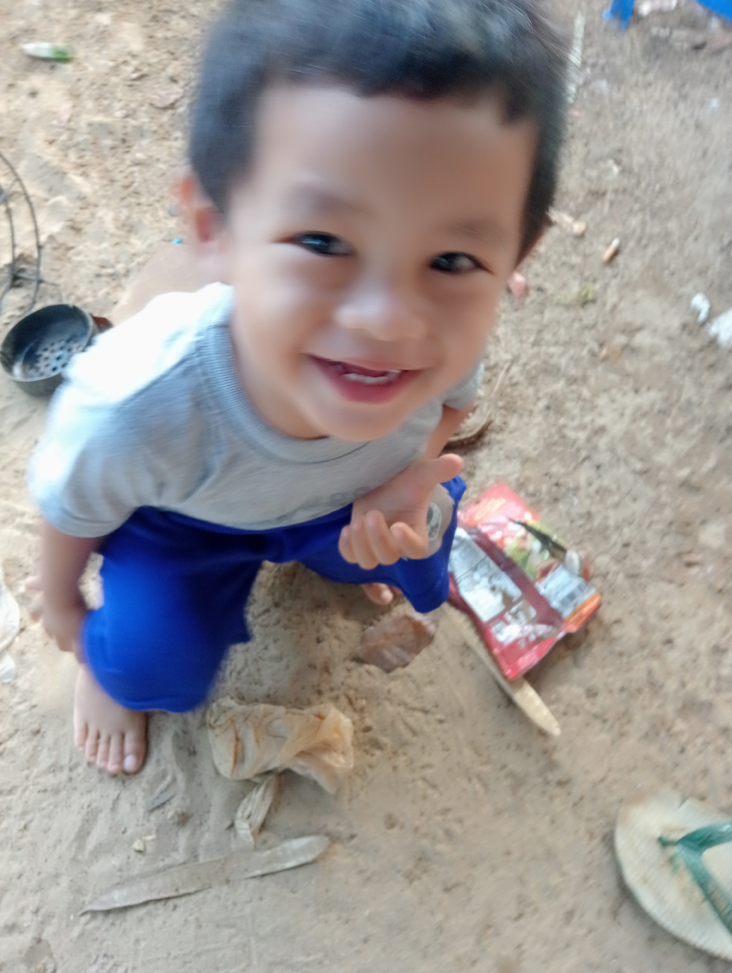 A young boy with dark hair and an open mouth, smiling, kneels on sandy ground in what appears to be an outdoor setting. He is wearing a light gray t-shirt and bright blue pants. To his left, there's a dark, perforated object. In the foreground and to his right, a brightly colored package, possibly food or candy, lies on the ground next to a small pile of brown, crumpled material. A single flip-flop with a green strap is visible in the lower right corner. The ground is a mixture of sand and dirt with some scattered debris. This scene is likely located in Muara, Indonesia, given the context.