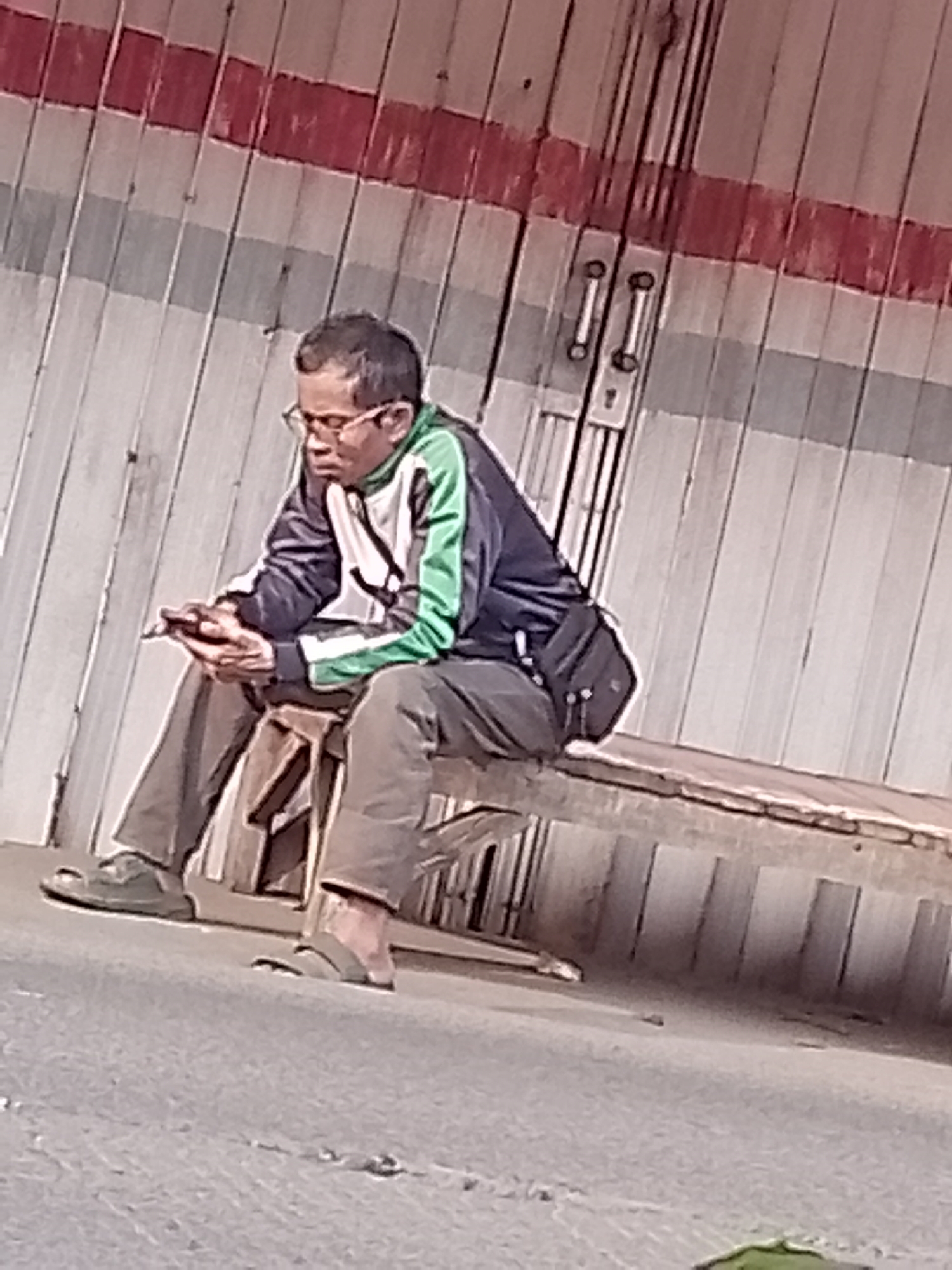 A man wearing glasses, a blue and green jacket, and brown pants sits on a wooden bench, looking at his phone. He is wearing black shoes on his left foot and sandals on his right foot. The bench is placed in front of a corrugated metal wall with red and gray stripes. The scene appears to be in an outdoor urban setting, possibly a bus stop or a public waiting area, in Cikalongkulon, Indonesia.