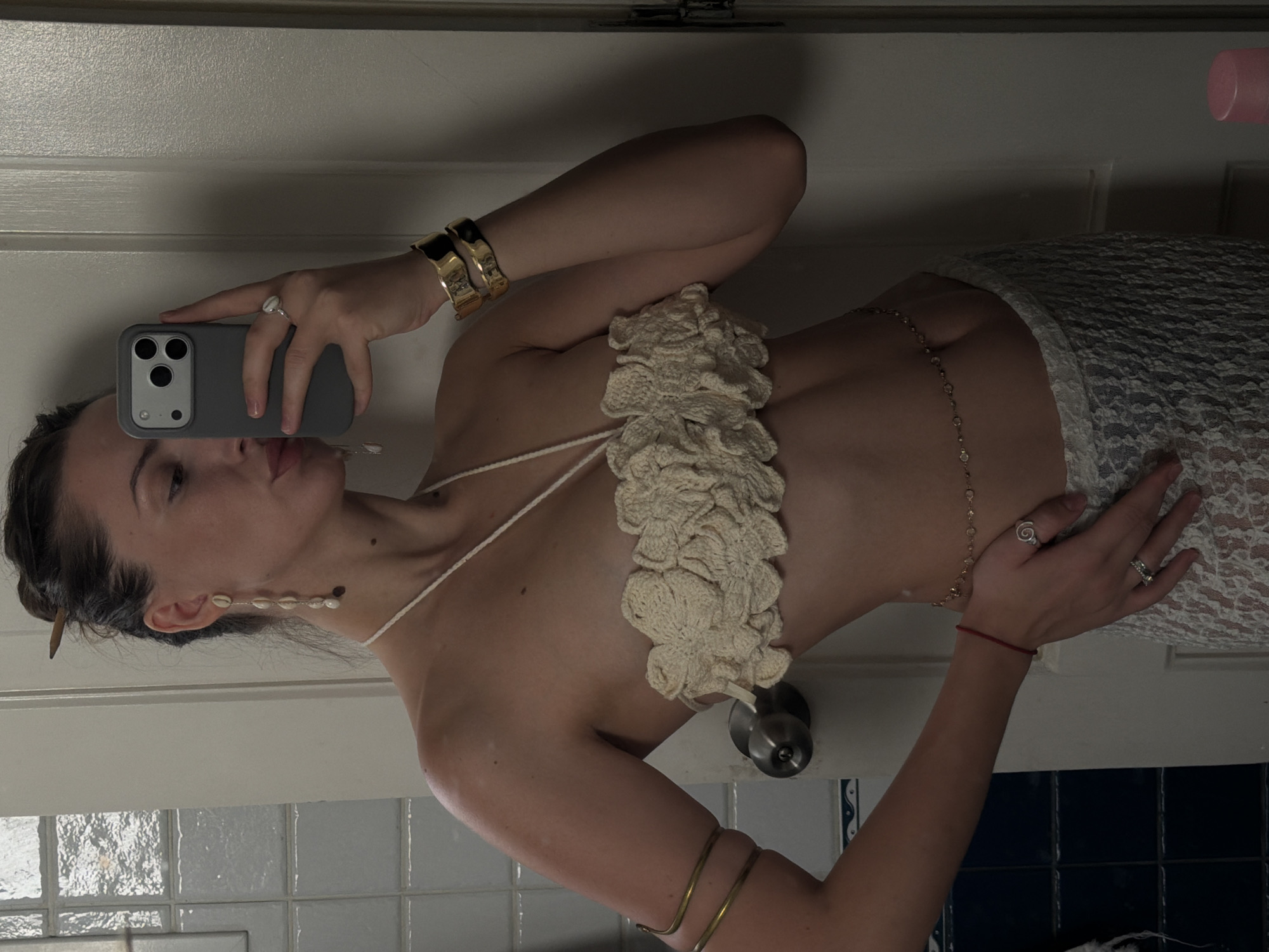 A woman stands in what appears to be a bathroom, taking a mirror selfie. She is wearing a white, crocheted halter top with floral detailing, a delicate gold belly chain, and a white lace skirt. She has long, dark hair styled in a bun, with a wooden stick holding it in place. Her accessories include a pair of long, dangling earrings made of small shells and pearls, a thin red bracelet on her left wrist, an arm cuff on her right arm, and two wide gold cuffs on her left wrist. She is also wearing two rings on her left hand, one with a silver spiral design and another with a clear stone. The background shows a tiled wall and a white door. The image does not provide any information or context related to Sharm El Sheikh, Egypt.