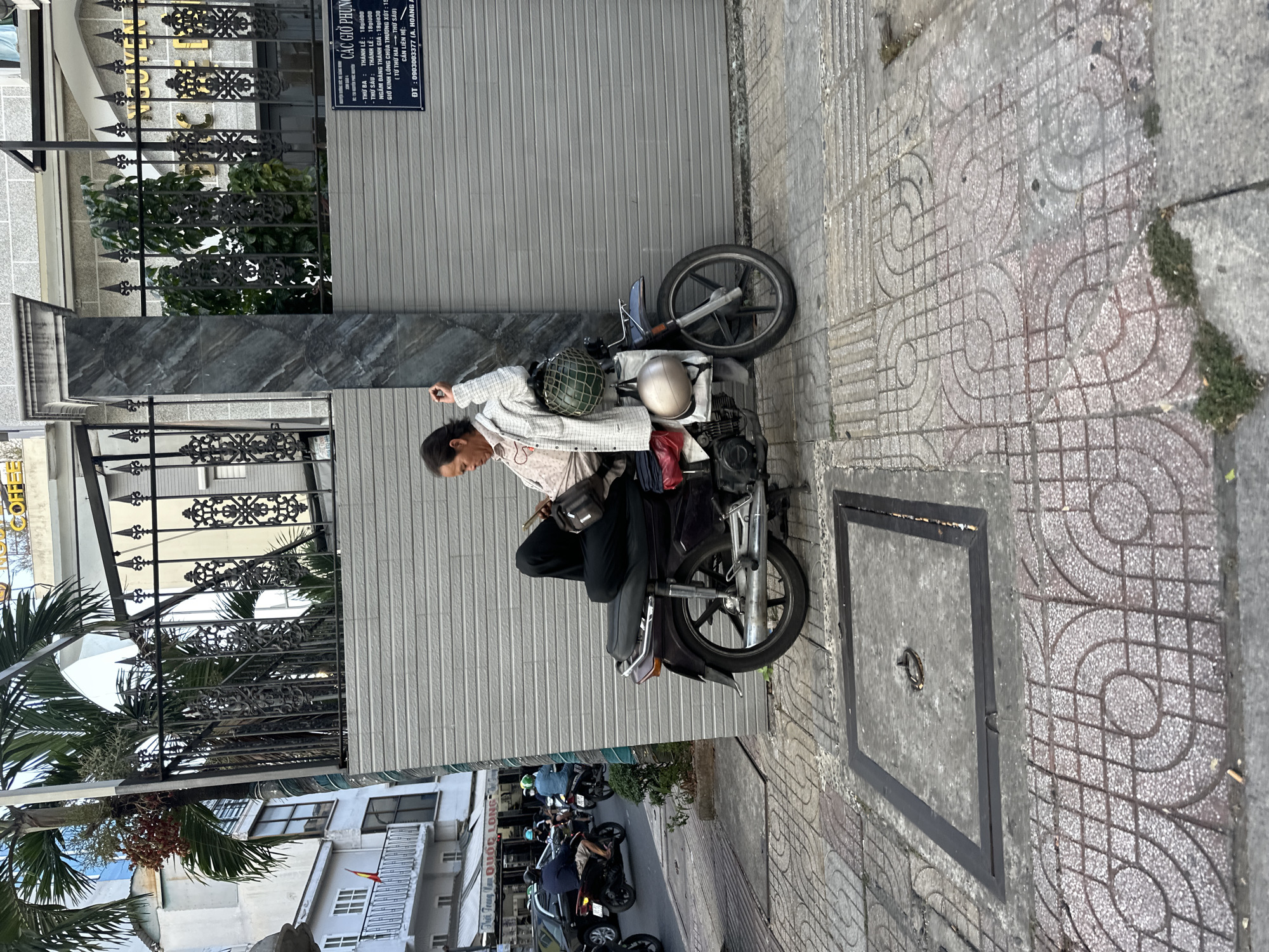 A middle-aged Asian man in a light-colored jacket sits on a parked moped in Thủ Đức, Vietnam. He is wearing dark pants and has a dark bag slung over his shoulder. The moped is parked on a patterned sidewalk next to a building with a gray corrugated metal wall. There are two helmets on the back of the moped. A manhole cover is visible in the sidewalk. Across the street, motorbikes are parked along the curb. A sign in Vietnamese can be seen on the building next to the moped. Palm trees and other buildings can be seen in the background.