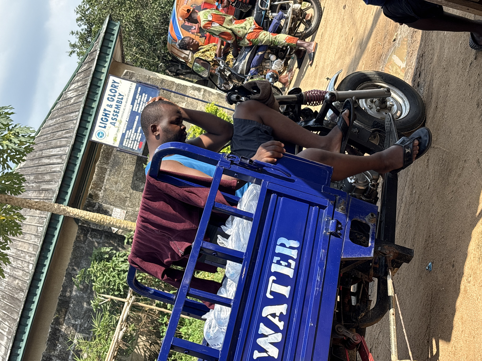A medium shot captures a man resting his head on his hand, sitting on a blue tricycle with "WATER" printed on its side. The tricycle appears to be filled with plastic bags of water. Behind him, a motorcycle carries two people, one dressed in a vibrant patterned outfit. The background shows a building with a sign that reads "LIGHT & GLORY ASSEMBLY, IKOT OFF ABANG ROAD BY CHRISTIAN SEC SOH ABAAPO IKOT ESER I EL." Vegetation and a clear sky can also be seen. The location is identified as Ikot Ekpene, Nigeria.