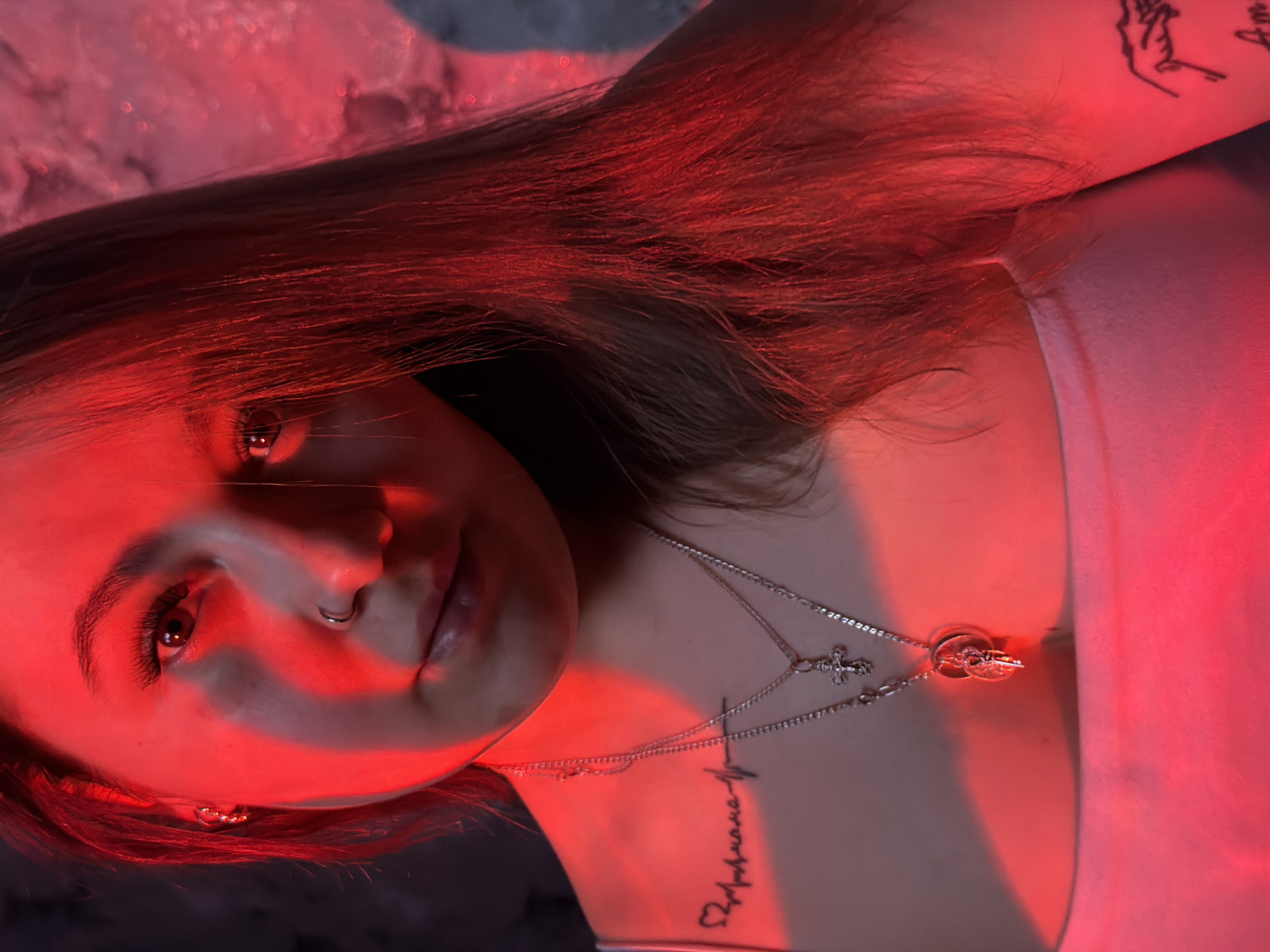 A young woman is prominently featured in a high-angle, close-up photograph, bathed in striking red artificial light that creates a dramatic and moody atmosphere. She is positioned with her head tilted, looking directly into the camera. Her long, dark hair, appearing reddish-brown under the intense illumination, frames her face and spreads around her.
Her visible features include a silver ring piercing in her left nostril and slightly parted lips. Her eyes reflect the strong red light, maintaining a direct and contemplative gaze. Adorning her neck are two delicate silver necklaces: one with a small cross pendant and another with a circular medallion.
Distinctive tattoos are visible on her body. On her right forearm, an abstract design depicts intertwined lines resembling two hands. A tattoo on her upper chest, near her collarbone, shows a heart symbol connected to a line reminiscent of an electrocardiogram (EKG), with the name "Ариана" (Ariana) written in cursive Cyrillic script below it. The background is softly blurred with deep red and dark grey tones, indicating an indoor setting. The composition focuses on the woman's expressive gaze and personal adornments, enhanced by the vibrant red lighting.