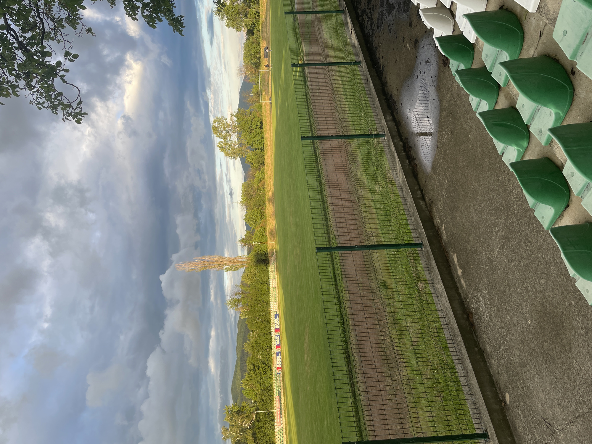 The image captures an empty outdoor sports field in Ihtiman, Bulgaria, viewed from the perspective of the spectator stands. In the foreground, on the right, rows of green and white plastic stadium seats are arranged on concrete steps. Below the seating area, a concrete drainage ditch shows standing water and reflections, suggesting recent rain. A green mesh fence separates the spectator area from the playing field.
The midground features a large, well-maintained green grass field, likely a football or soccer pitch, with a white goal post visible towards the left. Along the far side of the field, a colorful advertising hoarding or billboard displays repeating white "8888" figures on a red background, interspersed with other indistinct logos or graphics. Beyond the field, a line of green trees marks the perimeter, with a prominent tall, slender tree standing out on the left side.
In the background, distant mountains are visible beneath a dramatic and heavily clouded sky. The sky, occupying a significant portion of the upper image, shows a mix of dark, heavy clouds and lighter, illuminated patches, indicating late afternoon or early evening light. Tree branches with green leaves partially frame the top left corner of the sky.
The scene is tranquil and devoid of people or activity, conveying a quiet, post-event or pre-event atmosphere, possibly after a rain shower given the wet surfaces.