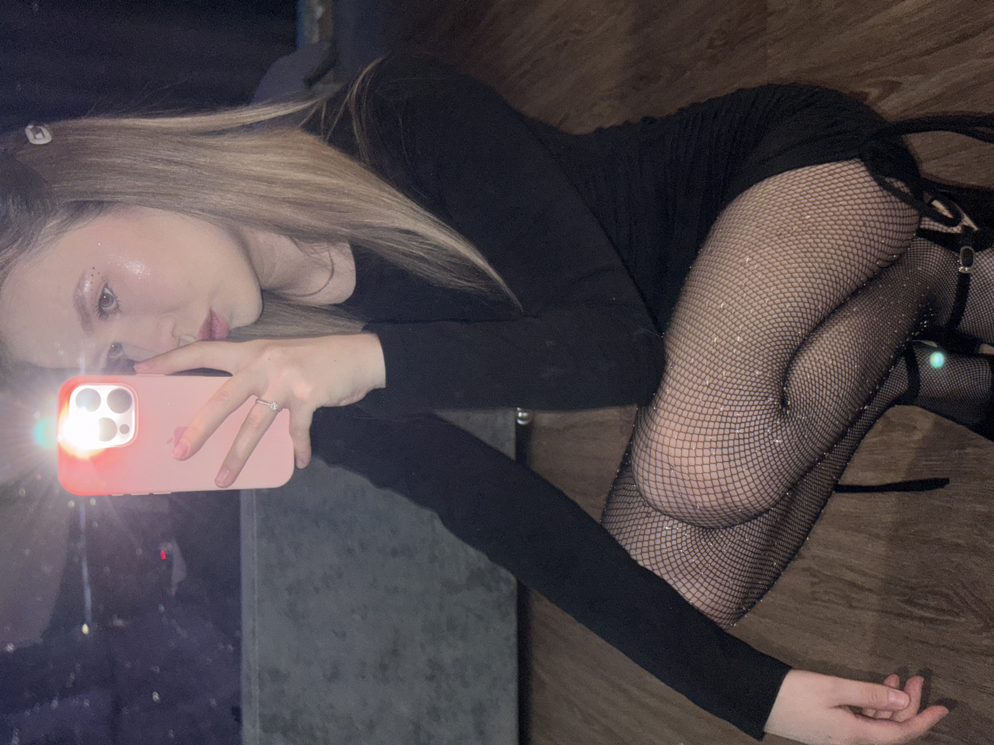 A young woman with long, light brown hair and fair skin is depicted lying on her side indoors in Chaikovsky, Russia. She is wearing a black long-sleeved top or dress and sparkly fishnet stockings, with a black strappy high heel partially visible on her right foot. A small white hair clip is in her hair, and a silver ring is visible on her left ring finger. She holds a pink iPhone with its bright flash illuminated, pointing it towards the viewer, suggesting she is taking a selfie or being photographed. Her gaze is directed towards the camera, with a neutral yet direct expression, and shimmering makeup highlights her left cheekbone. She lies on a dark wooden floor. To her left, a dark, reflective surface, possibly a window, reflects the phone's bright flash and some distant, indistinct lights, indicating the scene takes place at night or in a very dimly lit environment. No discernible text is visible.