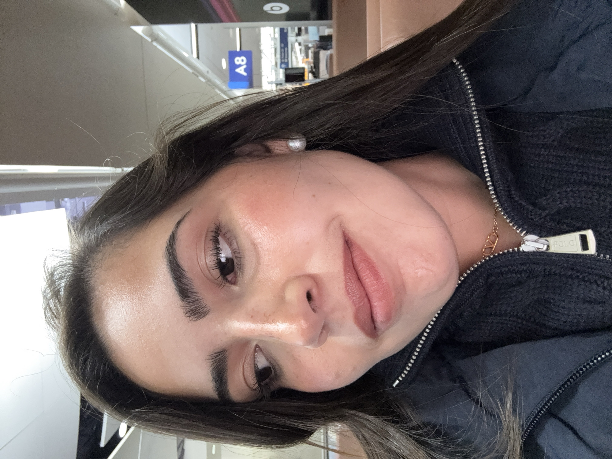 A close-up, angled portrait of a woman with dark hair and light skin. She is wearing a dark-colored jacket with a zipper. In the background, partially visible, is signage, including a blue sign with "A8" visible. This suggests the location might be an airport terminal. The city of San Antonio, United States of America, is not specifically discernible from the image.
