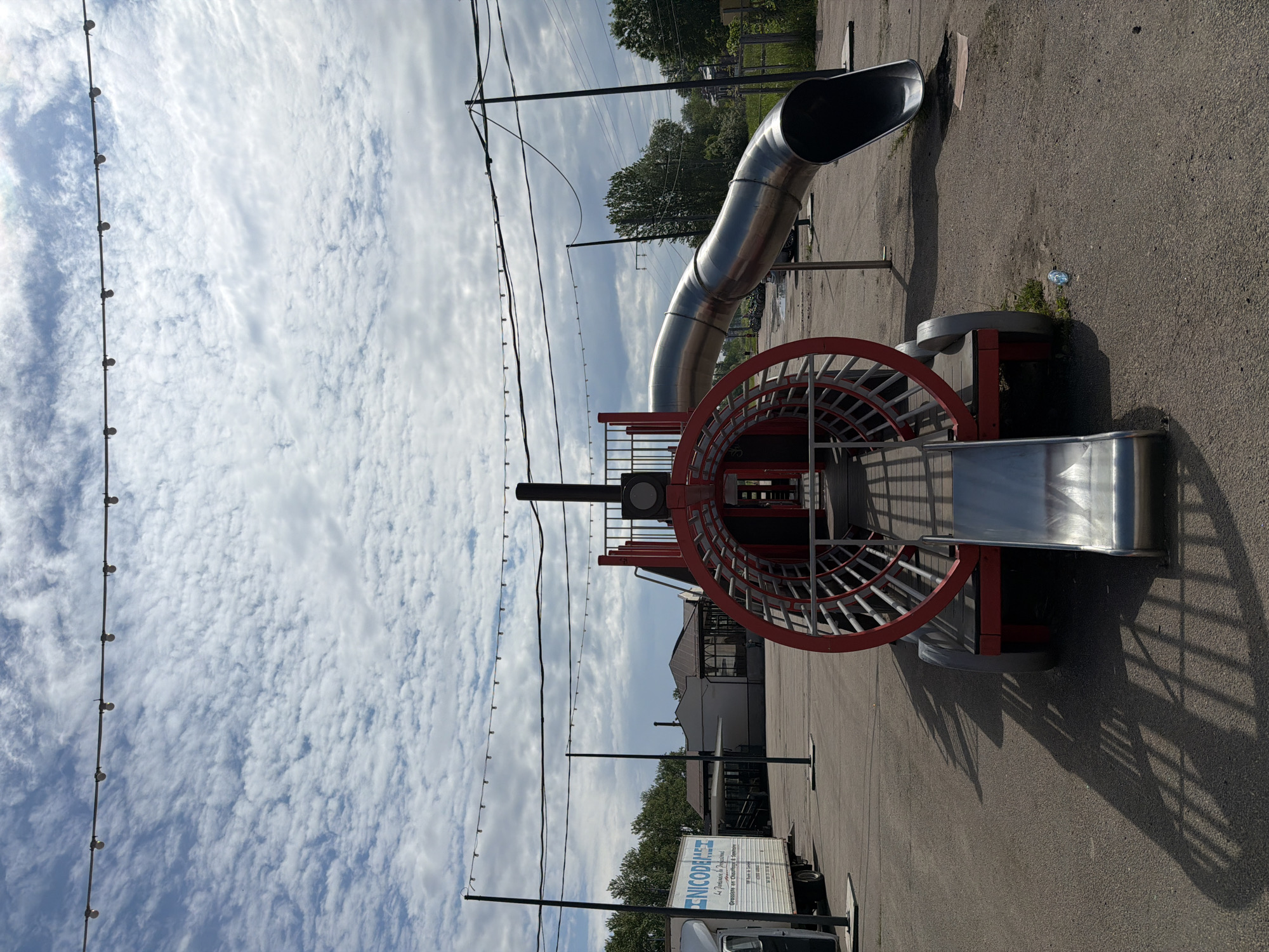 The image displays an outdoor scene centered around a children's playground structure. The main feature is a large, red, open-work cylindrical climbing apparatus with a flat top, from which two slides descend. One is a long, curved, enclosed metallic tube slide, while the other is a shorter, open, straight metallic slide. The ground beneath the playground is paved with asphalt.
No people are visible in the image, but the presence of the playground suggests a recreational area intended for children's play.
The scene appears to be set during daytime, under a bright, partly cloudy sky featuring extensive scattered cumulus or stratocumulus clouds. Soft shadows are cast by the playground equipment, indicating sunlight, possibly in the late morning or afternoon. Overhead, strings of decorative light bulbs are strung across the area, supported by dark poles, suggesting the space might also be used for evening events or gatherings. In the background, green trees are visible, along with indistinct buildings. A small, clear plastic bottle can be seen on the ground near the base of the playground. A white delivery truck is partially visible on the left side of the frame.
Text visible on the side of the white delivery truck includes the company name "NICODELEST" in blue letters. Below this, smaller French text reads "La Pâtisserie et Confiserie" (The Pastry and Confectionery), "Grossiste en Chocolats & Bonbons" (Wholesaler in Chocolates & Candies), followed by an address "108 Rue de la Glacière, 95260 BEAUMONT-SUR-OISE," and contact numbers "Tél. 01 39 31 22 93" and "Fax. 01 39 31 22 94."