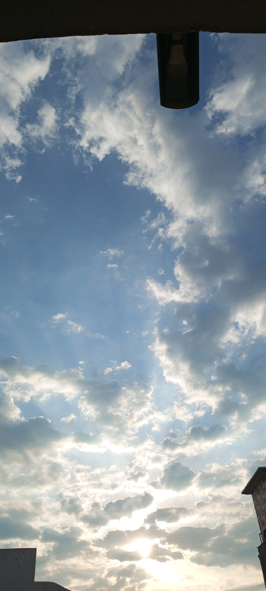 The image shows a view of a cloudy sky from a low angle, looking upwards. The top of the frame is partially obscured by a dark, horizontal structure, likely an overhang or roof. A cylindrical object, possibly a lamp or light fixture, hangs from this structure in the upper center of the image, with a lightbulb visible inside. The sky is a bright blue, filled with a variety of clouds ranging from wispy white to darker, more dense formations. Sunlight is breaking through the clouds, creating rays of light and illuminating parts of the sky. In the lower right corner of the image, a portion of a building with a brick pattern is visible. In the lower left corner, a dark, flat surface, likely a rooftop or ledge, is present. The overall scene depicts an outdoor urban environment with a focus on the sky.