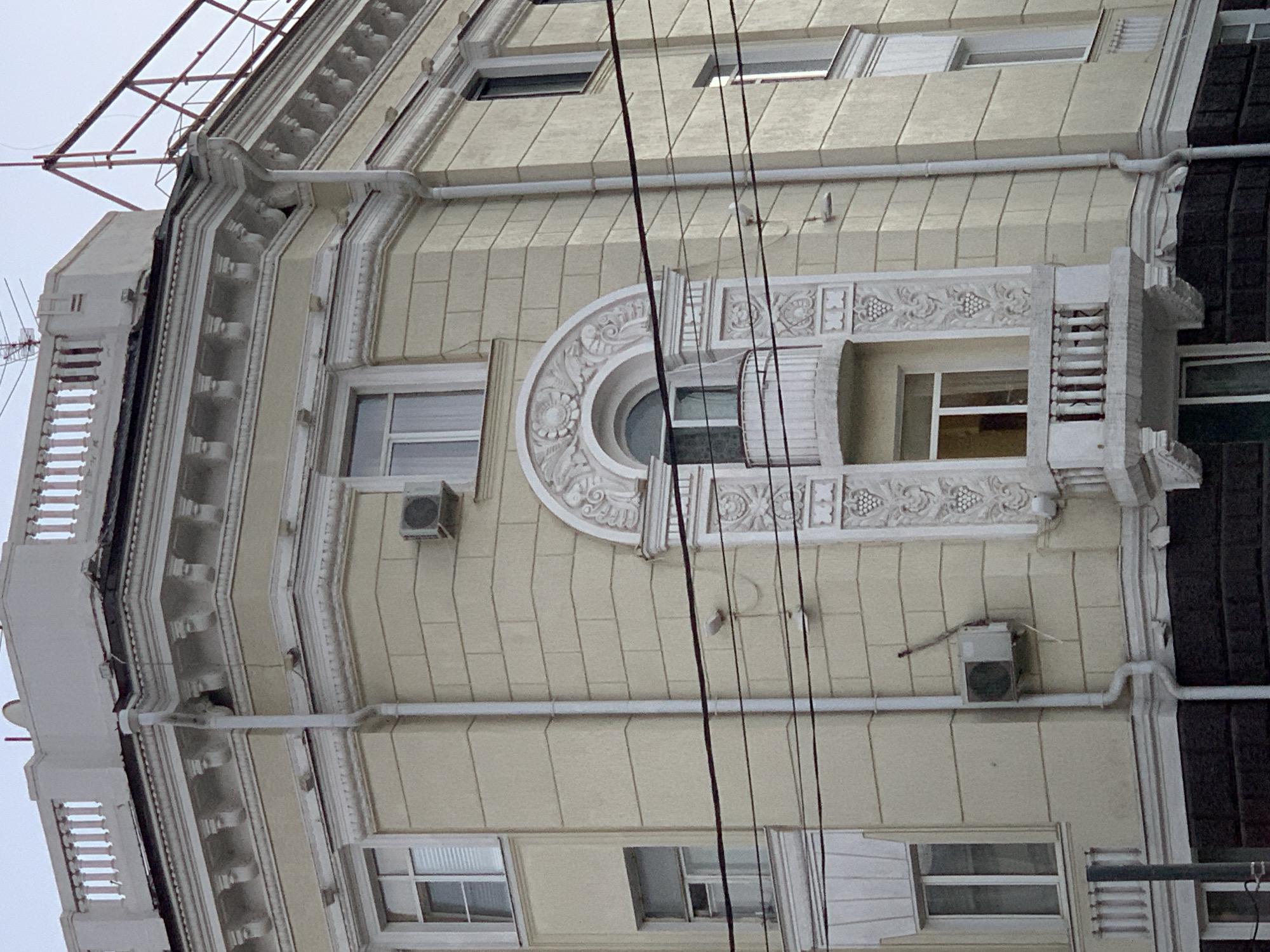 A light yellow, multi-story building with ornate architectural details dominates the frame. The building has a dark, textured base. Multiple windows, some with blinds, are visible across the facade. A white downspout runs vertically along the building's exterior. An air conditioning unit is mounted on the wall to the left of a rounded, decorative architectural feature that includes a small balcony with a white railing. Overhead, multiple dark wires are strung across the image. The sky is overcast and pale.