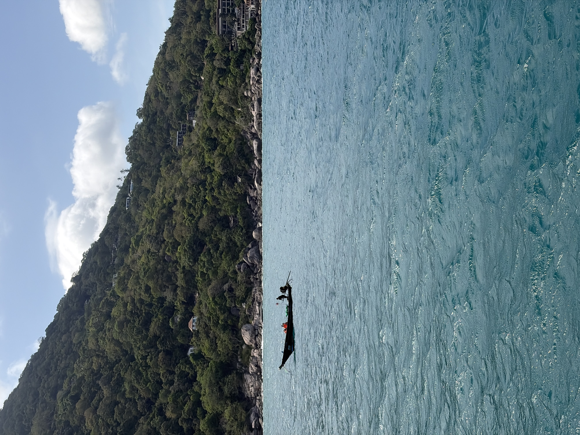 An outdoor, eye-level, horizontal medium shot shows a small, dark-colored boat with three people on it in the turquoise water of the Gulf of Thailand, off the coast of Ko Pha-ngan District, Thailand. The boat is in the left half of the frame, with the hull partially submerged in the wavy water. Two people are visible in the rear of the boat, and one person is in the front. The people appear to be wearing life vests. The boat has a number "66" painted on its side. In the background, a densely forested, steep hill slopes down to the water. Scattered along the hillside are several modern white buildings and structures. The shoreline is characterized by large, smooth grey boulders and rocks. The sky above is blue with scattered white clouds. The scene captures a tranquil coastal landscape.