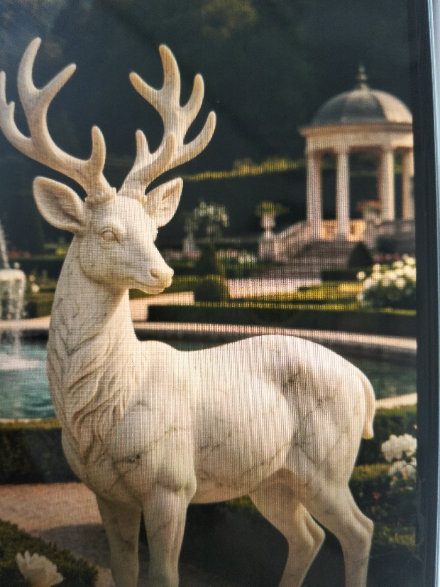 The image depicts a detailed marble statue of a stag in the foreground, facing to the right. The stag has large, branching antlers and is positioned in what appears to be a garden setting. In the background, a classical rotunda with columns and a domed roof is visible, along with manicured hedges, a fountain, and a body of water. The overall scene suggests a serene and ornate outdoor space, possibly a public garden or a private estate. The city of Payagaboh, Indonesia, is not depicted in this image; the architectural style and landscape are distinctly European in appearance.