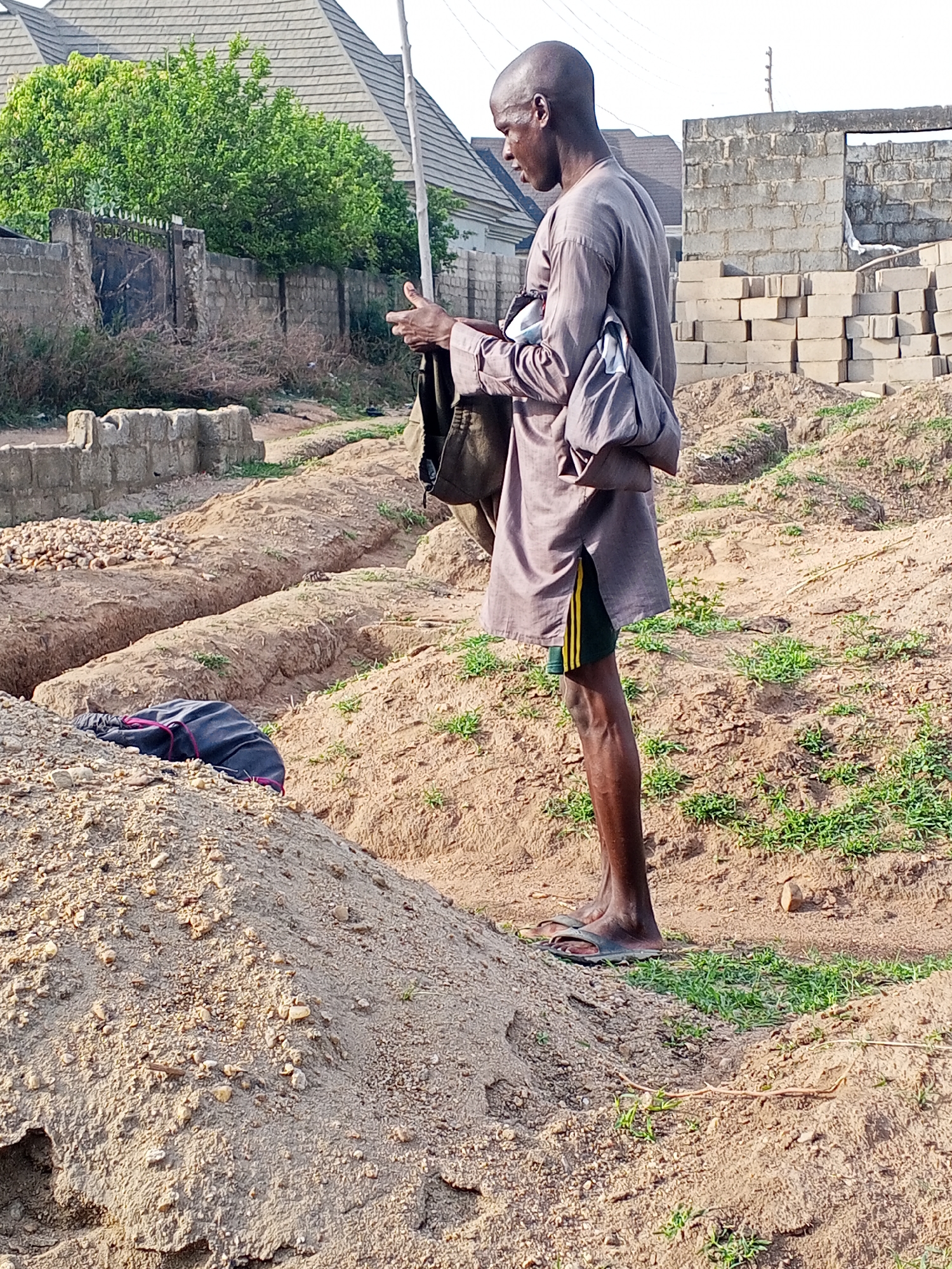 A man stands on a construction site near Zango-ndaji, Nigeria. He is light-skinned with a shaved head and is wearing a gray tunic, black shorts with yellow stripes, and flip-flops. He is looking down and holding something in his hands. A dark-colored bag hangs from his shoulder. In the background, there is a pile of building blocks, a partially constructed brick wall, and a building with a gray roof. The ground is sandy, and there are patches of green grass. A ditch is dug in the ground to the left. A dark-colored bag sits on a pile of dirt.