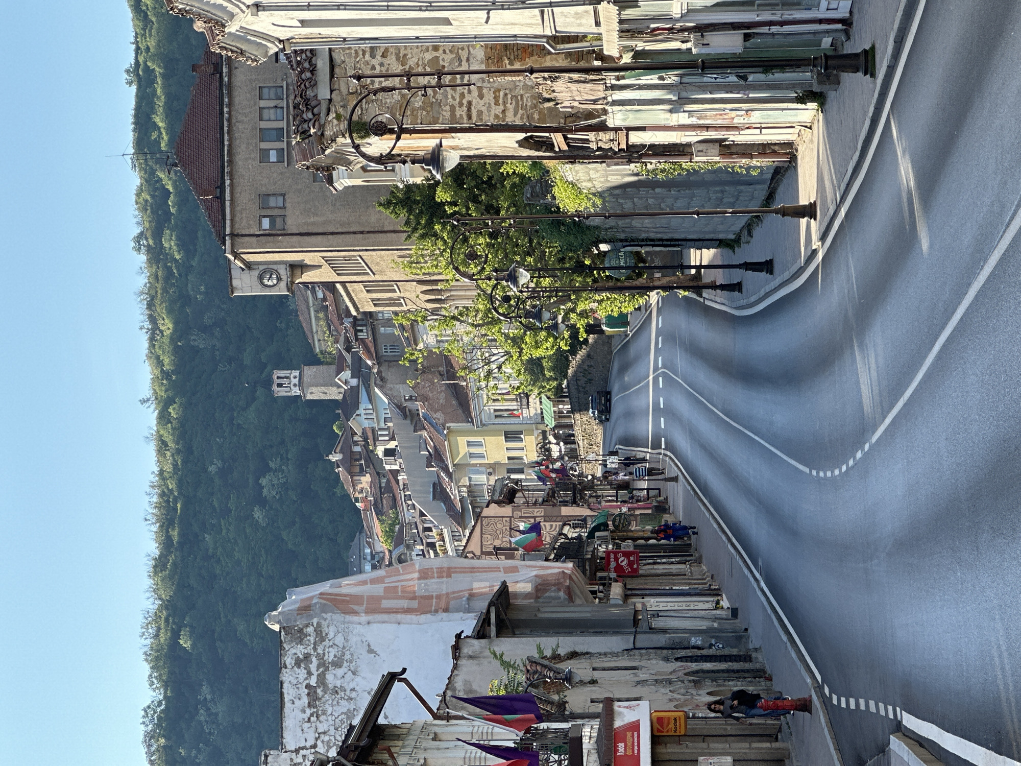 The image captures a street scene in Veliko Tarnovo, Bulgaria. A paved road winds through the old town, with white dashed lines marking lanes. Buildings line the street, varying in height, architecture, and condition, some adorned with flags. Streetlights punctuate the roadside. Along the edge of the street and on elevated platforms, market stalls or displays are visible, showcasing various items and crafts. The backdrop features a densely vegetated hillside, meeting a clear, light blue sky. A person is visible standing on the edge of the market with the road.