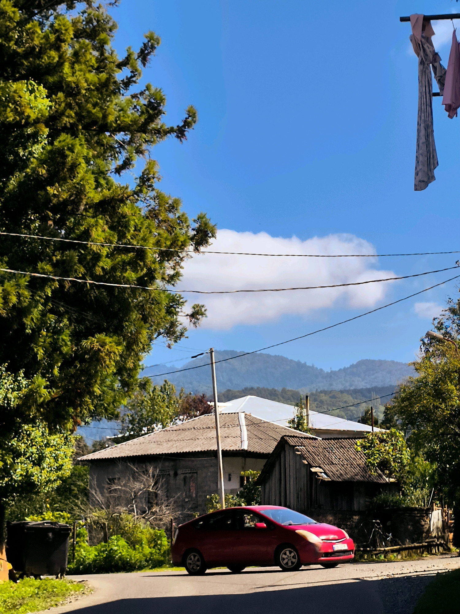 A red car drives on an asphalt road in front of a weathered grey house and a small wooden outbuilding. The car is a modern sedan, and its headlights are on. The house has a corrugated roof and several windows. The outbuilding is made of wood and has a partially collapsed roof. Lush green trees and vegetation surround the buildings. In the background, rolling hills are visible under a bright blue sky with scattered clouds. There are power lines strung across the scene. Laundry is hung on a line in the upper right corner of the image. The scene appears to be a rural setting, potentially in Erge, Georgia, given the architectural style and vegetation.