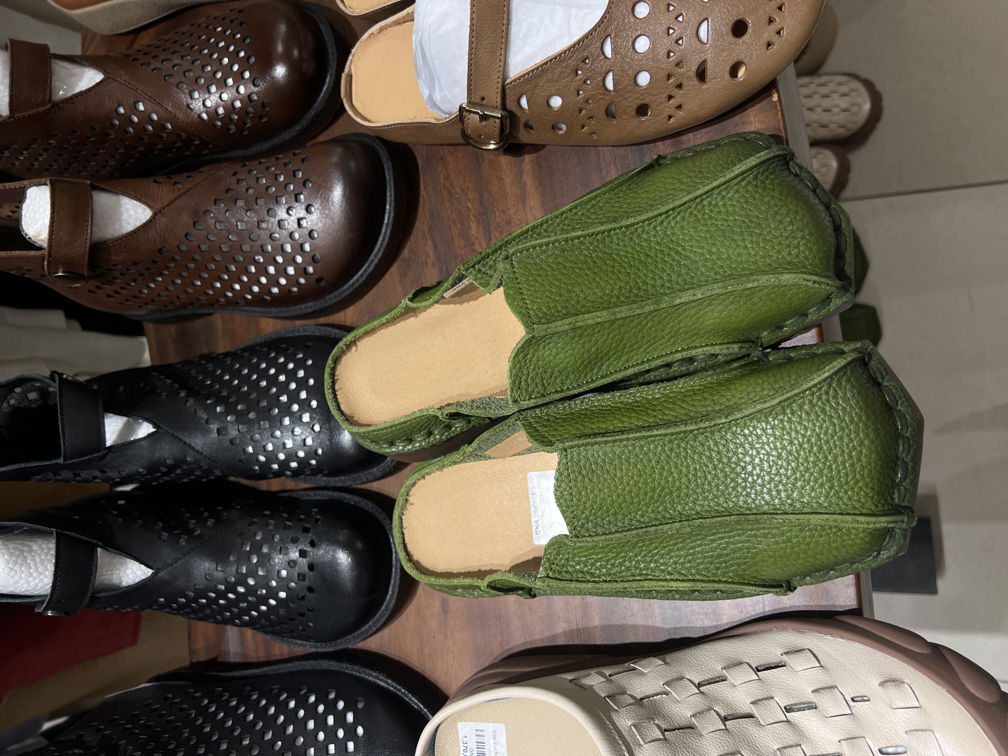 The image shows a display of various shoes on a wooden surface. The shoes include several pairs of brown and black leather shoes with perforated designs, a light brown sandal with cut-out patterns, a pair of green leather slip-on shoes, and a pair of woven light beige shoes with brown soles. The scene is possibly located in a shoe store in Nha Trang Ward, Vietnam, as indicated by price tags in Vietnamese currency visible on some of the shoes. The focus is on the arrangement of the shoes, showcasing different styles and colors available for purchase.