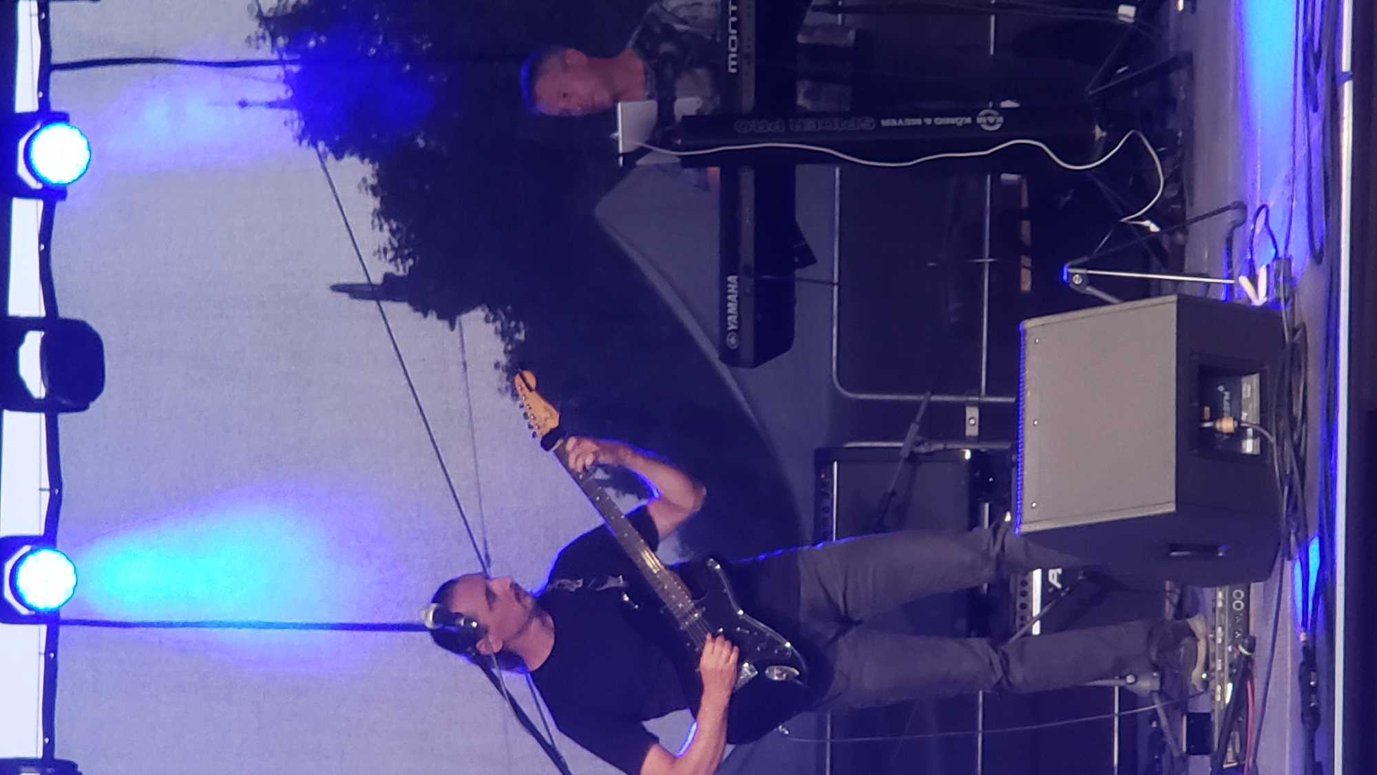 The image depicts a band performing on an outdoor stage at an event in Prešov, Slovakia. In the foreground, a guitarist with long dark hair tied back, wearing a black t-shirt and grey pants, is actively playing a black electric guitar. He is positioned on the left side of the frame, with his body turned towards the instrument. To his right, a keyboardist with short grey hair, wearing a grey and black patterned t-shirt, is seated and playing a black keyboard with a "YAMAHA" logo visible. The keyboardist is looking forward, towards the audience or performance. Behind the musicians, there are stage lights, a backdrop, and trees visible in the background. A large black speaker is placed on the stage in the foreground, to the left of the center. The lighting on stage is primarily blue and white, creating a vibrant atmosphere. The overall scene suggests a live music performance taking place at night.
