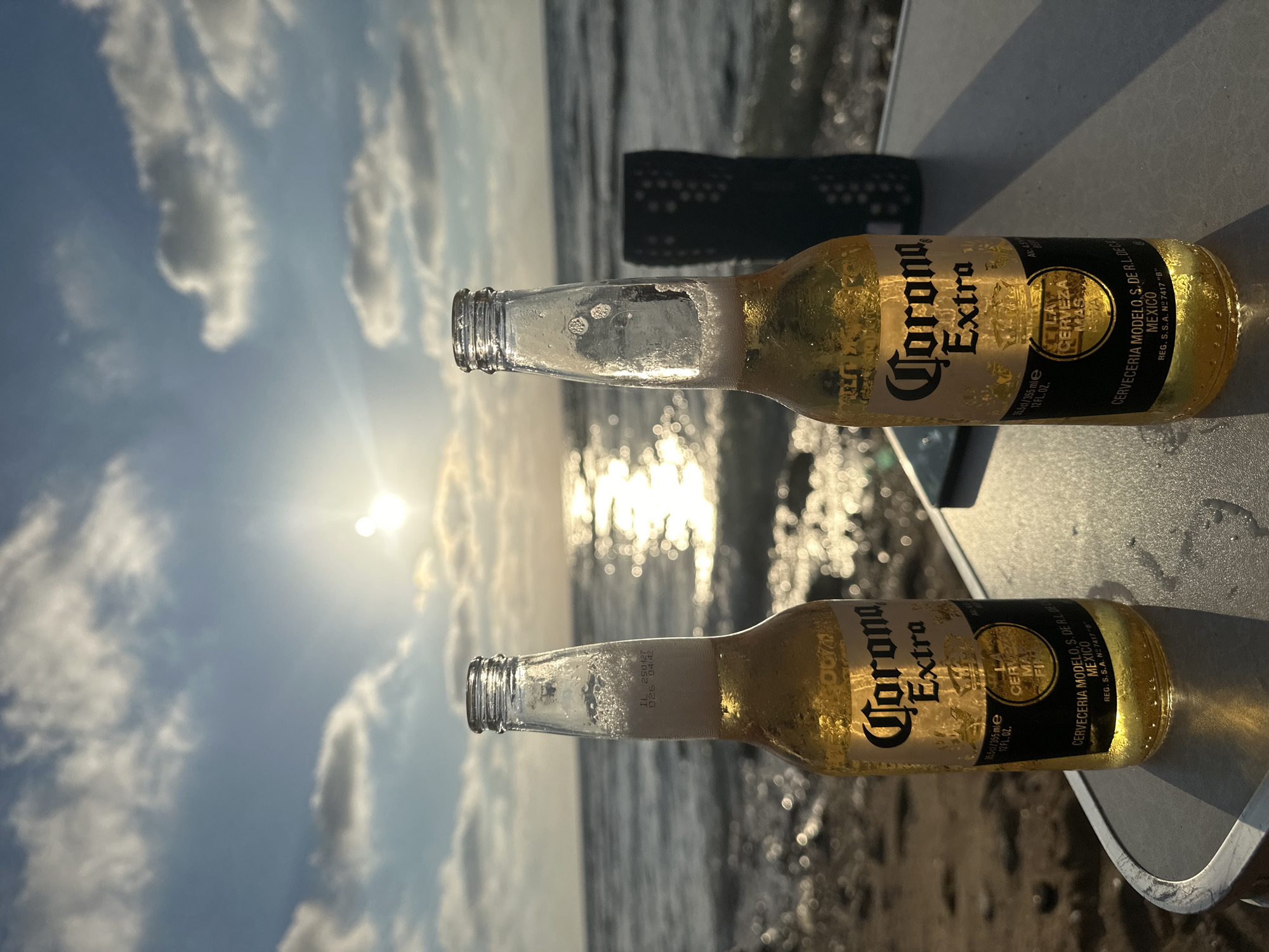 Two bottles of Corona Extra beer are on a table at a beach in Haifa, Israel, with the Mediterranean Sea in the background and the sun setting. The bottles are clear glass with yellow beer and white foam at the top. Both bottles have black and gold labels with the Corona Extra logo. The table is round and white, with some water droplets on its surface. To the right of the bottles, a dark green cup is visible. The beach sand is visible in the foreground, and the ocean water reflects the sunlight. The sky is a mix of blue and white clouds, with the sun low on the horizon, suggesting it is either sunrise or sunset. The overall scene depicts a relaxed setting by the sea.