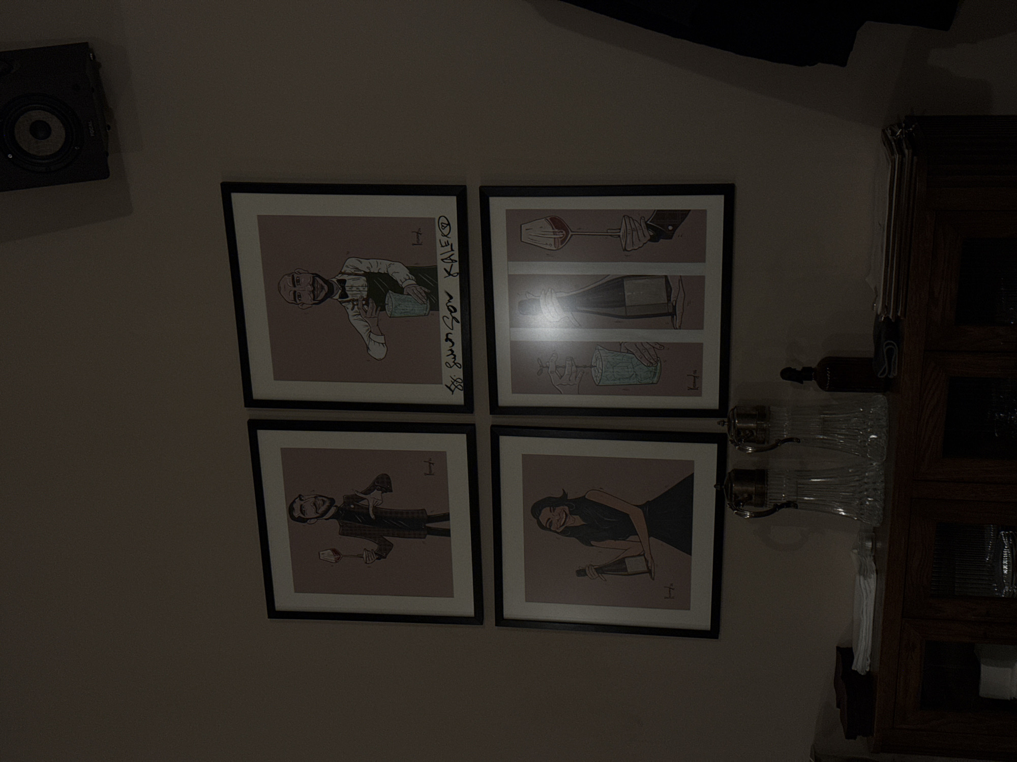 Four framed drawings hang in a square formation on a wall. A small black speaker is mounted above and to the left of the drawings. The drawings depict caricatures of people interacting with wine. Next to the drawings is a dark wooden cabinet with glassware. The image's location is most likely indoors, but its location within Yerevan, Armenia, cannot be determined.