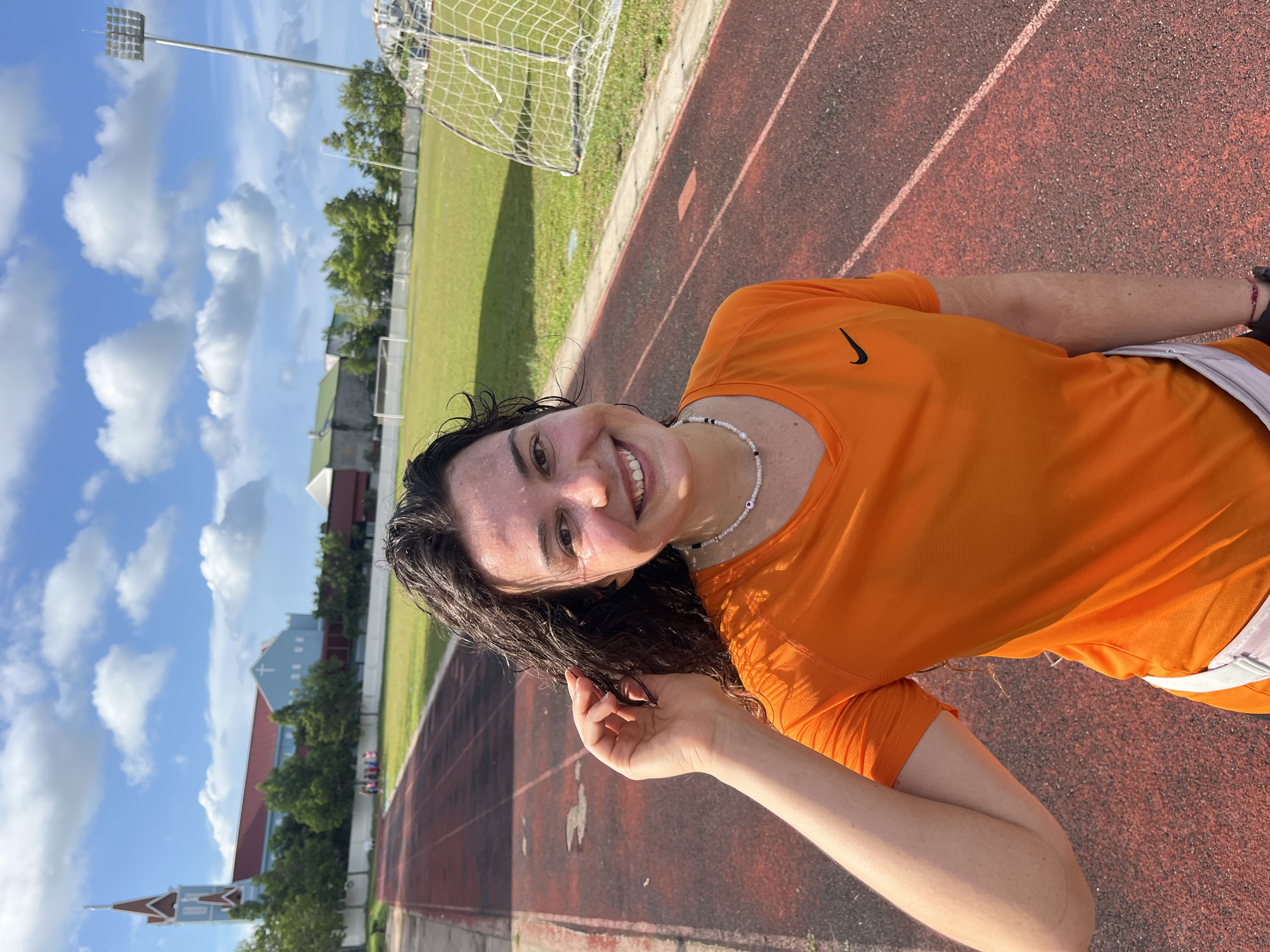 A young woman with dark, curly hair and fair skin is smiling at the camera, seemingly after a workout. She is wearing a bright orange athletic shirt with a black Nike swoosh logo and a white and black beaded necklace. Her face is glistening with sweat, suggesting she has been active. She is on a red running track, with a green grassy field and a soccer goal visible in the background. The sky is bright blue with fluffy white clouds, indicating it is a sunny day. In the distance, there are some buildings and trees. The overall mood of the image is cheerful and energetic.