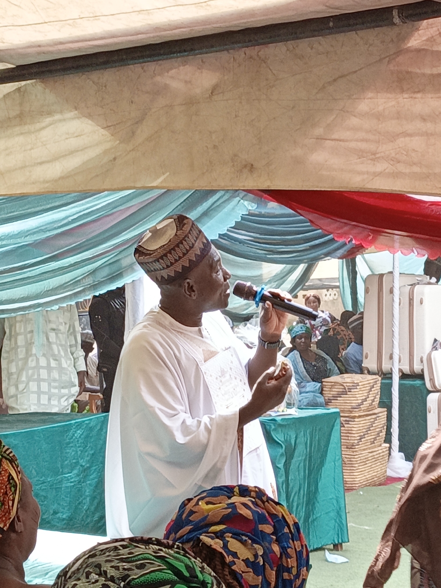 This image captures a daytime event or gathering, likely taking place outdoors under a large canopy or tent in Sabon Gari West, Nigeria. The overhead structure features a light-colored fabric canopy supported by dark poles, with decorative drapings of teal and red fabric underneath.
The central subject is an adult man, positioned mid-frame and facing right, speaking into a blue and black microphone held in his right hand. He is dressed in a traditional white long-sleeved garment, possibly an agbada, and wears a distinctive patterned cap (kufi) with geometric designs in brown and beige. In his left hand, he holds a small, white, unidentifiable object, possibly a card or small ornament. He appears focused on his address.
In the background, other individuals are partially visible. To the left, another person wears a white, patterned top. Towards the right, several cream-colored suitcases are stacked, and below them, multiple woven, rectangular baskets are neatly piled, suggesting gifts or ceremonial items. Further back, obscured figures include a woman in a blue outfit and headscarf. In the immediate foreground, the tops of heads of attendees, adorned with colorful patterned headwraps, are visible. A table covered with a teal cloth is also present, and a small piece of white paper rests on the green ground covering.
The scene suggests a formal or ceremonial occasion, such as a celebration, public address, or religious event, with the speaker taking a prominent role. The bright lighting indicates it is daytime. No discernible text is visible on any objects.