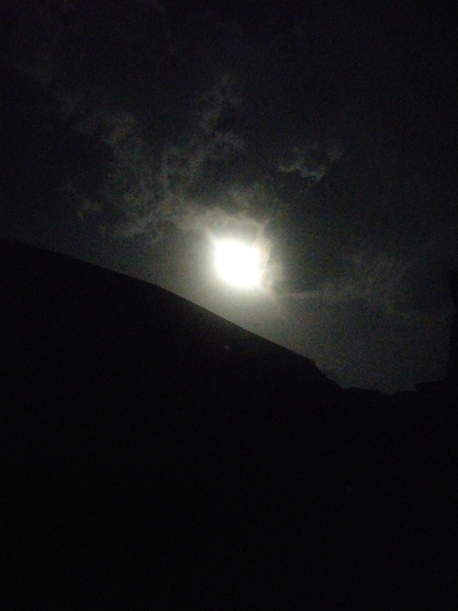 The image shows a nighttime scene with a bright light source, likely the moon, obscured by clouds. A dark, silhouetted object, possibly a building or hill, occupies the lower portion of the frame. The overall environment appears dark and indistinct. The location is Unknown, Unknown. No people or subjects are visible.