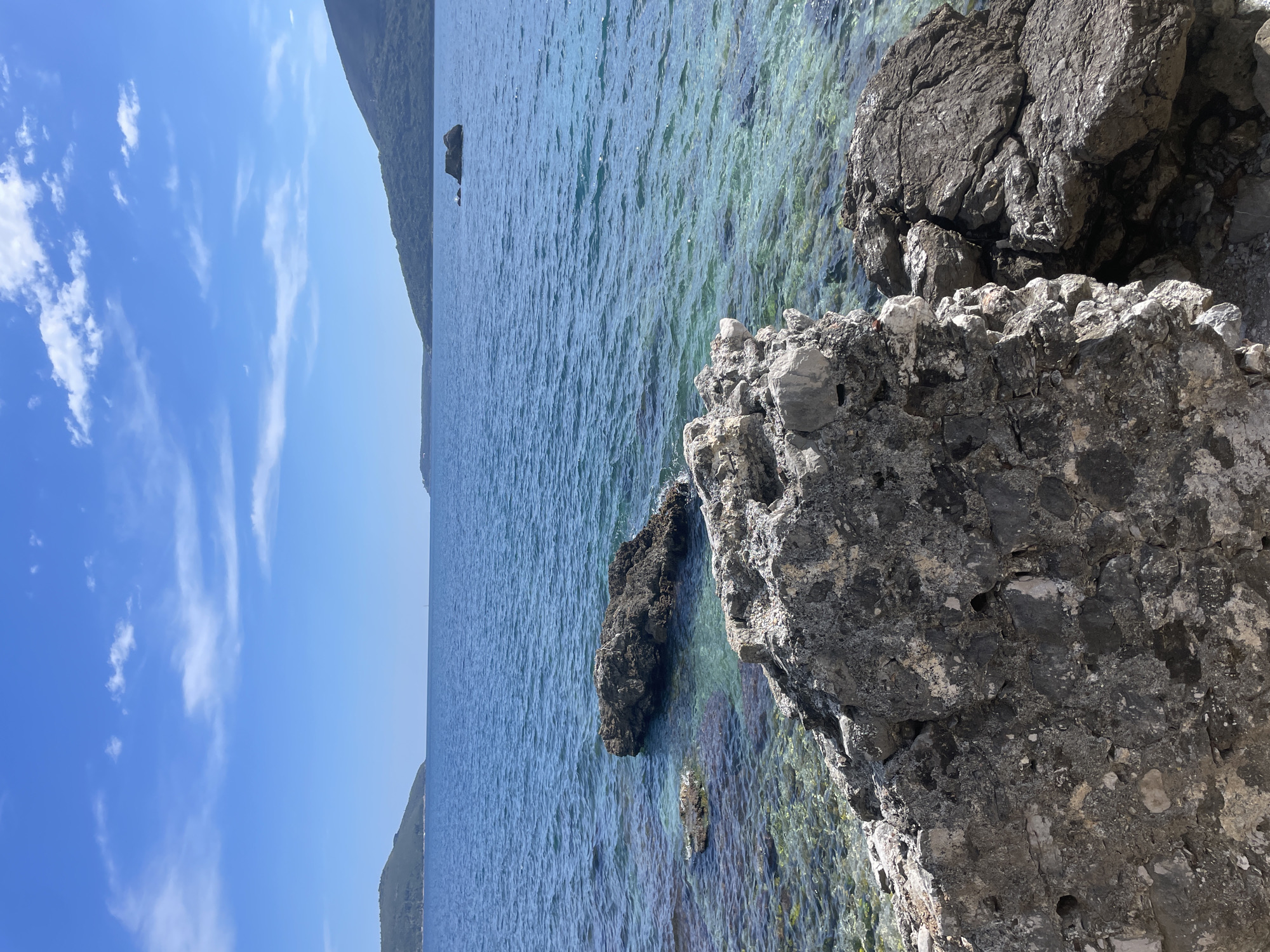 The image shows a seaside view in Herceg Novi, Montenegro. The Adriatic Sea is visible, with clear, blue-green water. Large, gray rocks form the shoreline in the foreground. A distant mountain range is present. The sky is bright blue with wispy clouds.