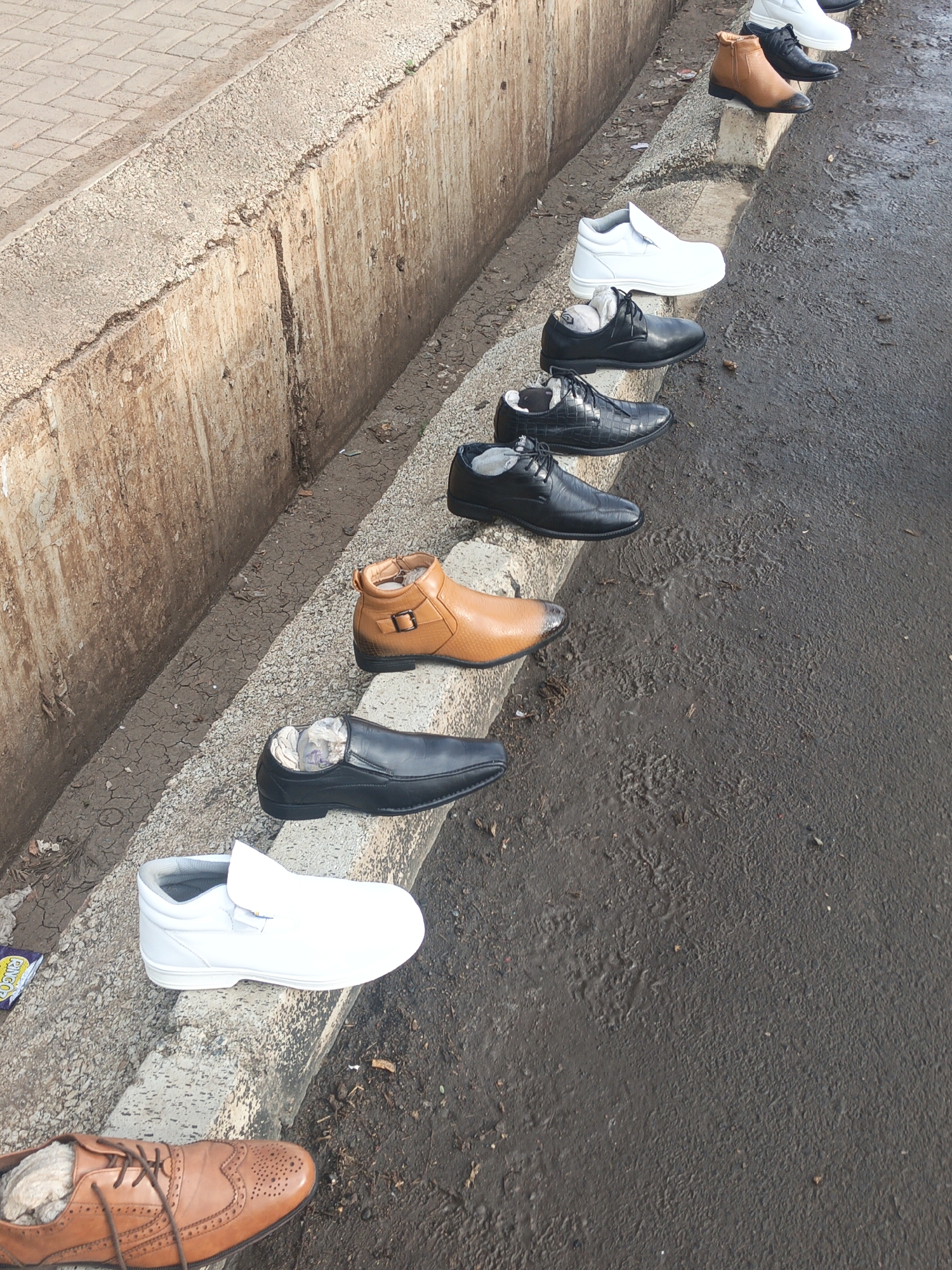 The image shows a row of shoes lined up on a curb alongside a street in what appears to be Nairobi, Kenya. The location is urban, characterized by a concrete curb and a street with signs of wear and possible moisture. The footwear consists of various styles, including dress shoes and casual boots, in colors like black, brown, and white. Some shoes have paper inserts inside. The placement suggests they are possibly for sale or display along the roadside. The curb itself has a weathered appearance, with signs of dirt and discoloration. The street appears wet or has a residue that gives it a darker tone. The setting is a typical street scene, conveying a sense of everyday commerce. There are no people visible in the photo.