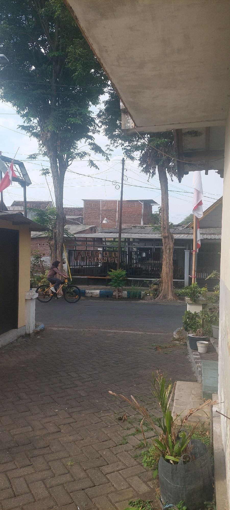 This image depicts a street scene in Lumajang, Indonesia, with a person riding a bicycle. The foreground is a paved walkway made of dark gray interlocking bricks. A person wearing a brown headscarf and a brown hooded shirt is riding a yellow bicycle with black tires on the asphalt road. The person is positioned in the left half of the frame, moving towards the right.
Behind the road, there is a dark metal fence with the word "KEDAI" visible in white block letters. Beyond the fence, there are several buildings with tiled roofs, some appearing to be made of brick. Two large trees with green leaves stand prominently in the middle ground. One tree is to the left of the road and fence, and another is to the right, with its trunk visible.
In the background, more buildings and trees are visible under a hazy sky. Two Indonesian flags, one red and white, are displayed on poles. The overall environment suggests a residential or commercial street in Indonesia.