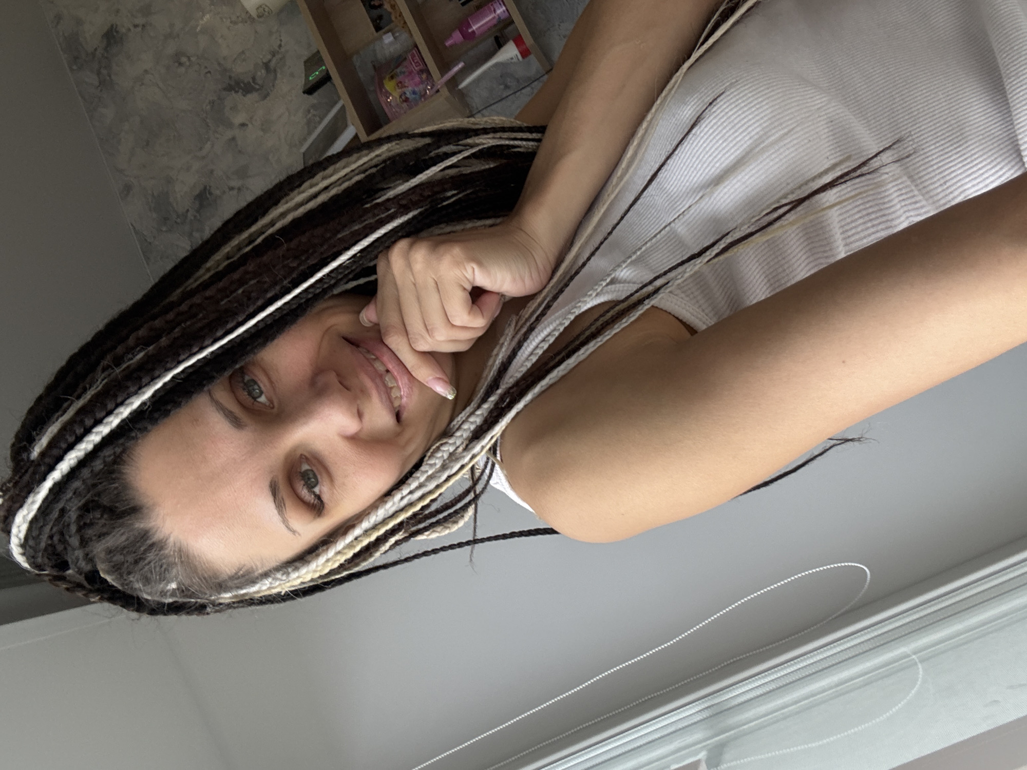 A portrait of a woman with her face at the lower-left corner of the frame. She has light skin, light eyes, and her hair is styled in black and white braids. She wears a light-colored, possibly white, tank top. Her right hand is touching her face, resting on her chin.
Behind the woman is a gray wall, and there is a shelf with various items on it in the upper-left corner. A window with white blinds is visible in the lower-right corner.
There is no location information provided, therefore the location in the city of Unknown, Unknown cannot be verified.
