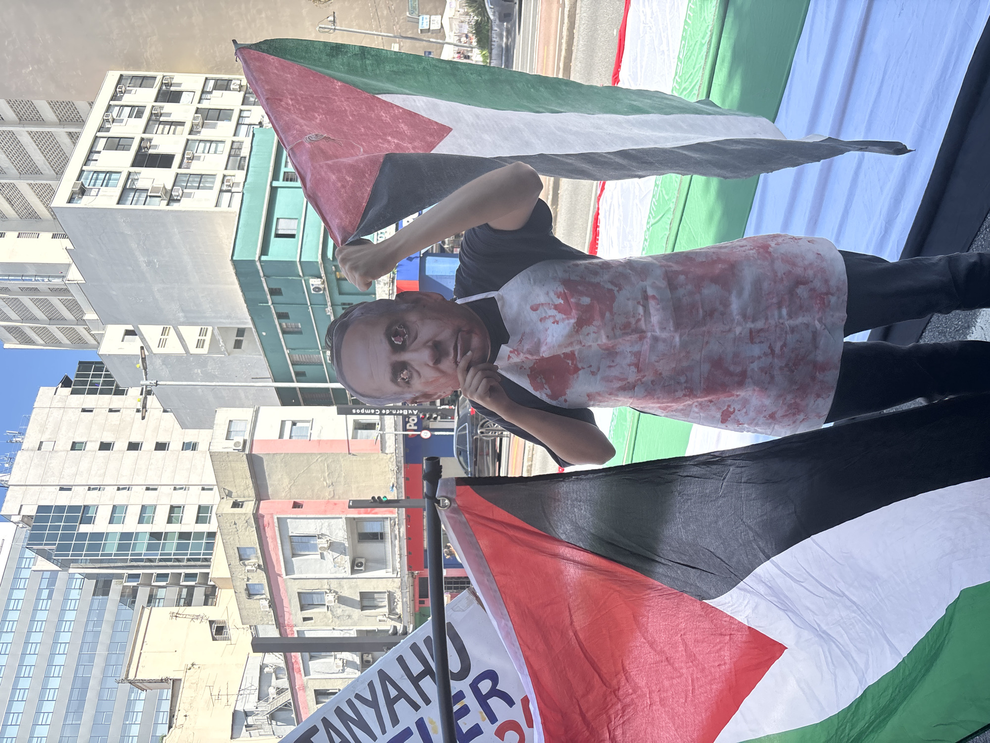 A protester stands in front of a backdrop composed of a Palestinian flag and another flag that reads "Netanyahu = Hitler." The individual wears a mask with the likeness of Israeli Prime Minister Benjamin Netanyahu, stained with what appears to be fake blood. They also wear a blood-spattered white apron. The location is identified by a sign reading "Av. Rebouças" to be in the city of São Paulo, Brazil. Buildings characteristic of São Paulo architecture surround the scene.