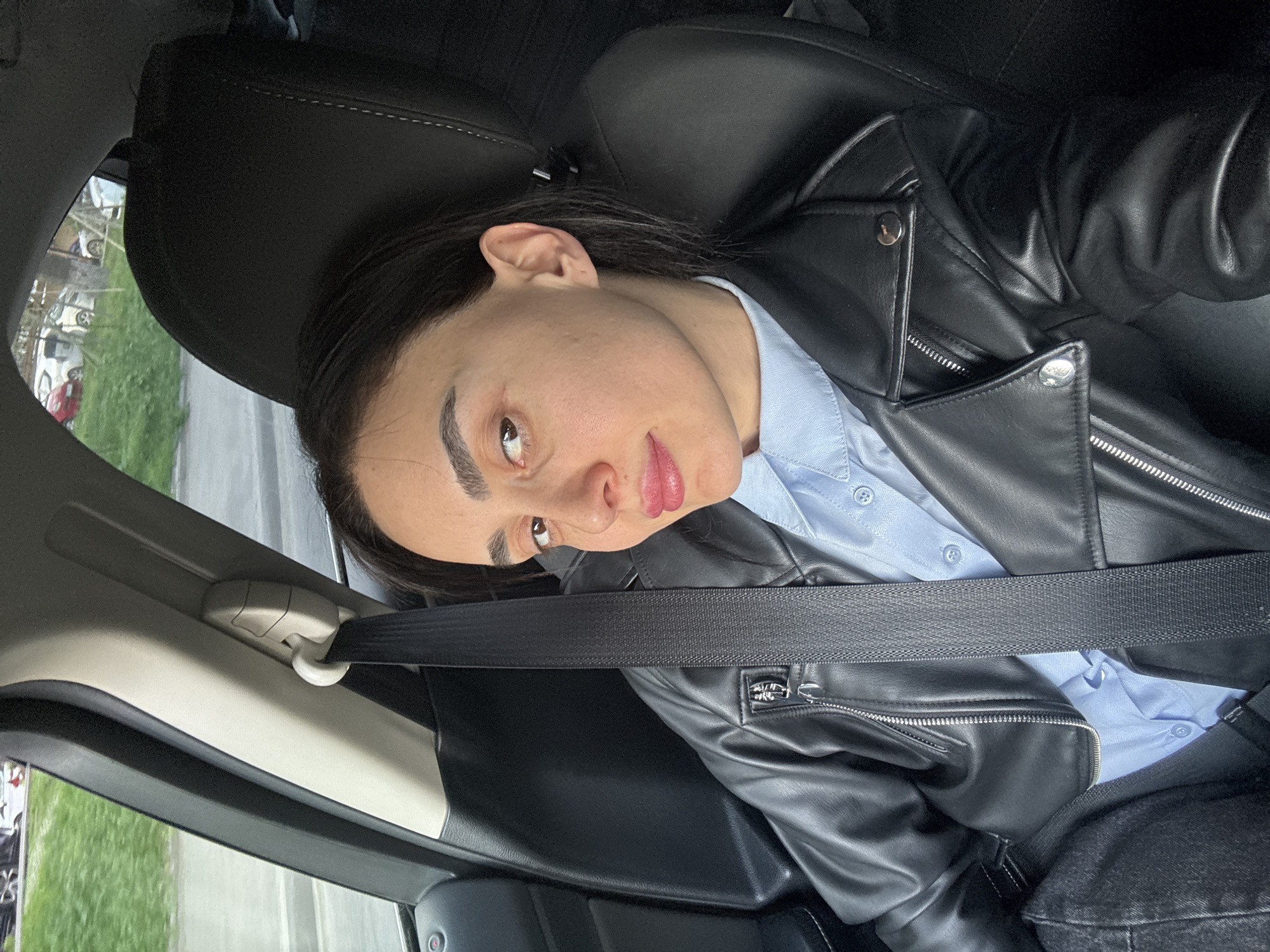 A person is seated in the passenger seat of a vehicle wearing a black leather jacket over a blue button-up shirt. A seatbelt is fastened across their chest. The environment outside of the vehicle's window suggests an urban area with other parked vehicles and some greenery. Based on the vehicle’s interior and the style of the surrounding environment, it is not possible to confirm this is the city of Tbilisi, Georgia. No specific actions or interactions are visible in the image.