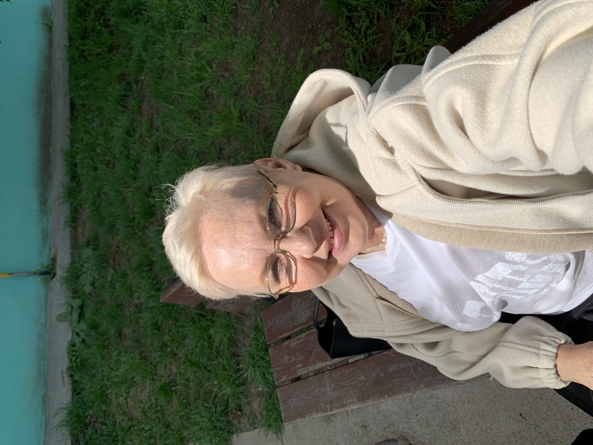 A mature woman with short blonde hair and glasses sits on a wooden park bench outdoors. She is wearing a white t-shirt and a light beige hoodie. The bench is situated in a green area with grass, and a building with a light blue facade is visible in the background. A black bag sits next to her on the bench. The scene is well-lit. No information indicates that the image was taken in Chaikovsky, Russia.