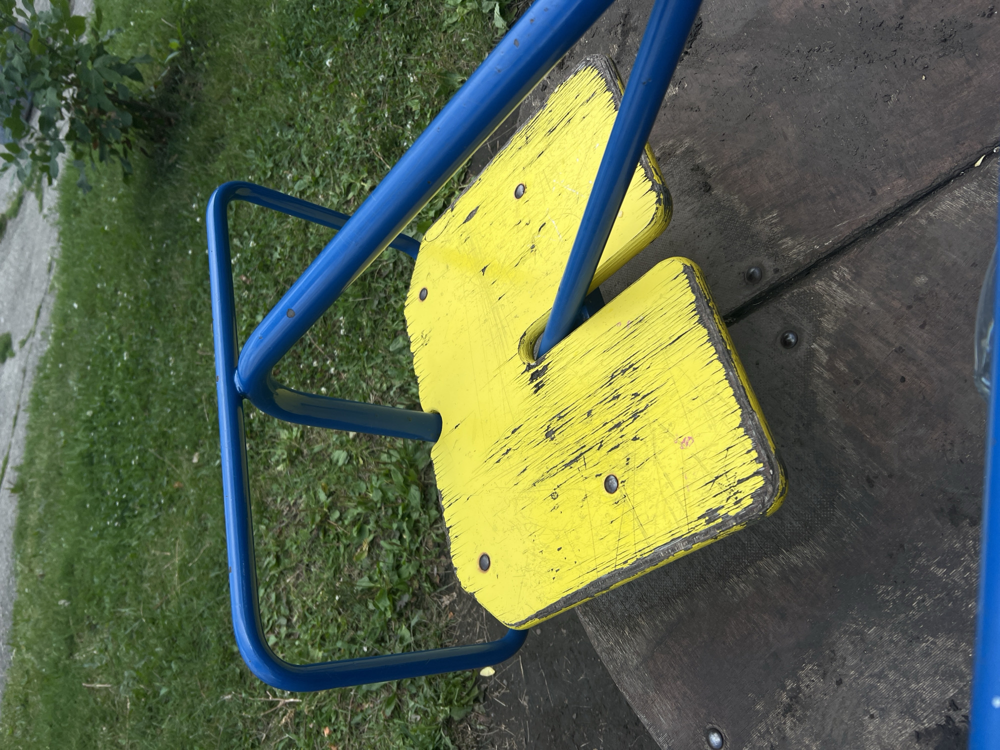 The image shows a close-up, angled view of a weathered yellow seesaw seat with blue metal supports, situated on a wooden platform. The background consists of green grass and some foliage. There are no people visible in the image. The scene appears to be outdoors, likely in a park or playground setting. The specific location in Ulyanovsk, Russia, cannot be determined from the visual information alone. The seesaw seat shows significant wear, with scratches and chipped paint, indicating it has been used extensively. The wooden platform beneath is dark and appears somewhat dirty. The lighting suggests it is daytime.