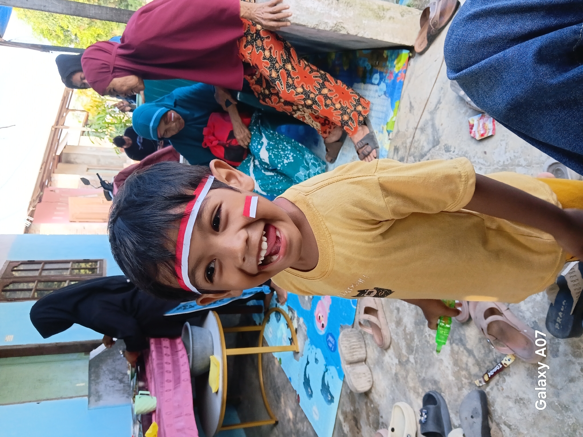 In Tembung, Indonesia, a young boy with face paint depicting the Indonesian flag smiles broadly, his mouth open in laughter, in the foreground of an outdoor scene. He wears a yellow t-shirt and has red and white stripes painted across his forehead and a small red and white flag sticker on his cheek. Behind him, several other individuals are visible. To the left, a person wearing a black hijab is partially visible, engaged in an activity near a blue wall and a table with pink cloth. In the mid-ground, a woman in a blue headscarf and another in a maroon hijab are seated. The woman in maroon wears patterned orange pants. A motorcycle is parked in the background. The ground is a concrete surface with a colorful mat depicting sea creatures. Various footwear, including sandals and flip-flops, are scattered around the area. The overall setting appears to be a residential outdoor space.