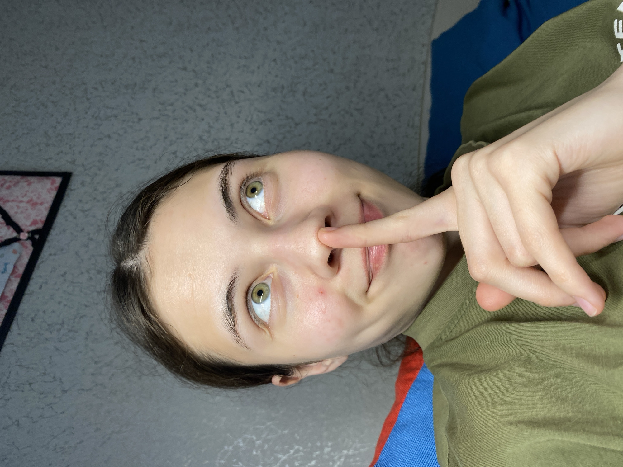 A young woman with fair skin, green eyes, and dark hair touches the tip of her nose with her index finger. She is wearing an olive-green shirt. The background includes a gray wall and a partially visible blue and red blanket or fabric. The location is indeterminable.