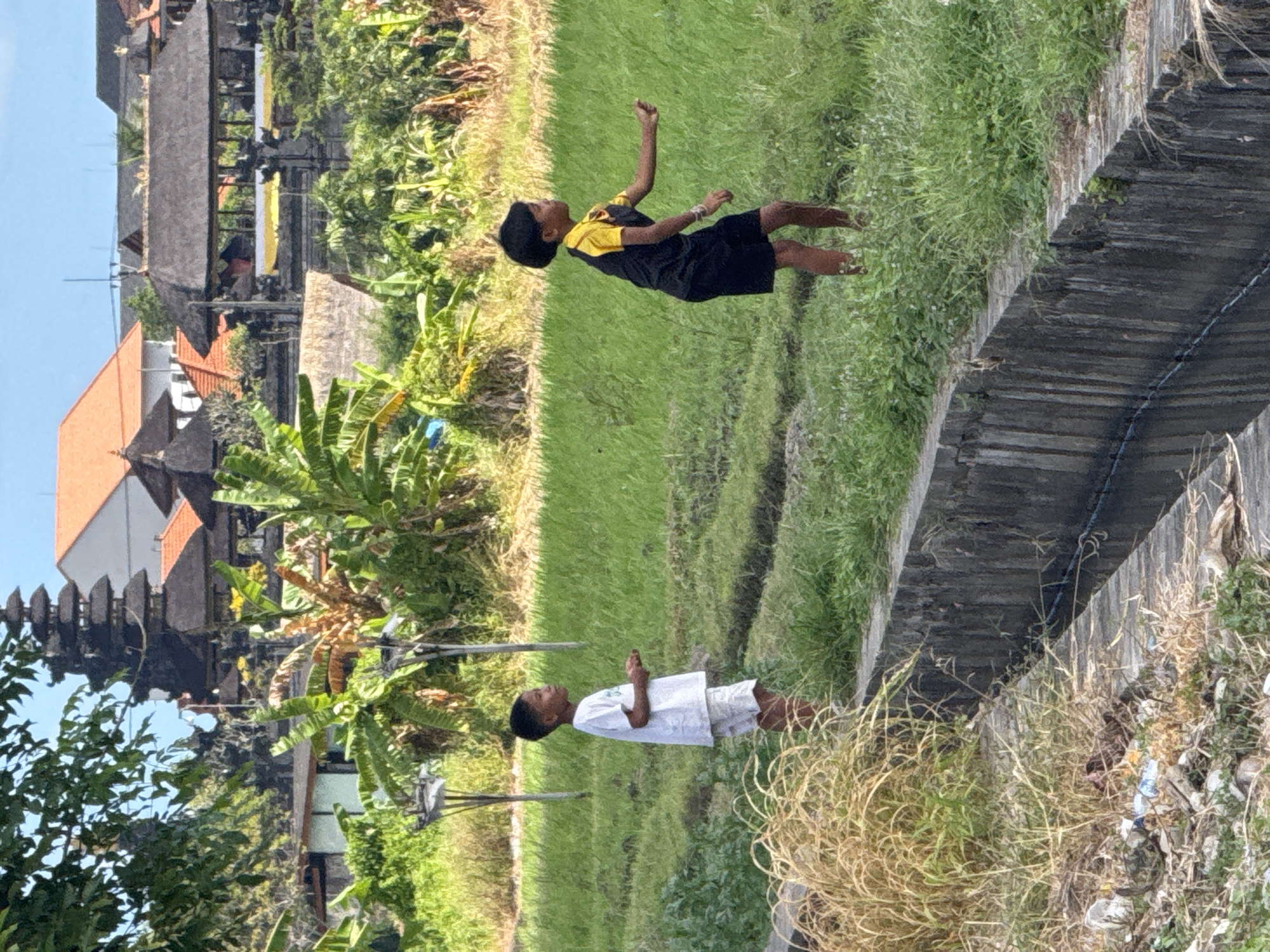 The image shows two children in a rural area. One child, wearing a black shirt with yellow sleeves and black shorts, is standing on a grassy area near a ditch. The other child is standing on another grassy area wearing a white t-shirt and white shorts. Behind them are rice paddies and a building. The location is likely Mengwi, Indonesia, based on the presence of rice paddies and traditional Balinese architecture.