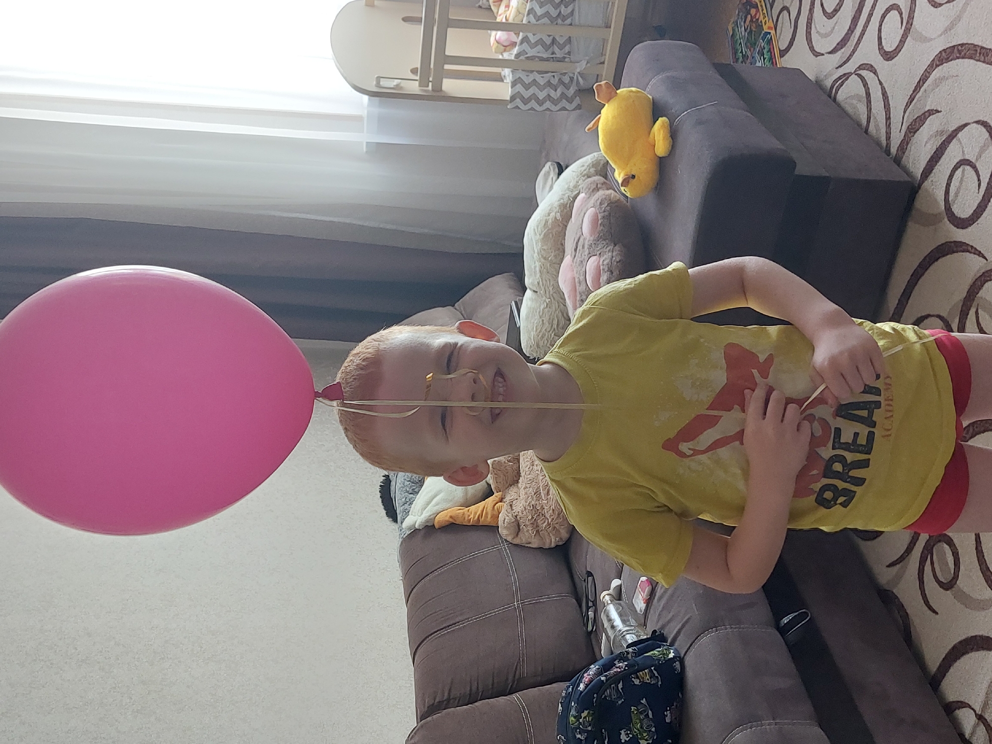 A young, fair-skinned, red-haired child wearing a yellow t-shirt with red trim on the sleeves and red shorts is positioned on a brown sofa in what appears to be a residential interior in Chaikovsky, Russia. The child is smiling and has a pink balloon with a string tied around their head. The balloon floats near the beige wall. The child holds a thin object in their hands. On the couch, there are several stuffed animals, including a yellow duck, a white bear, and others. A diaper-changing table stands in the background near a window with blinds. A patterned carpet with brown swirls lies on the floor. Other objects such as a water bottle and a backpack are visible on the couch. The scene is lit naturally.