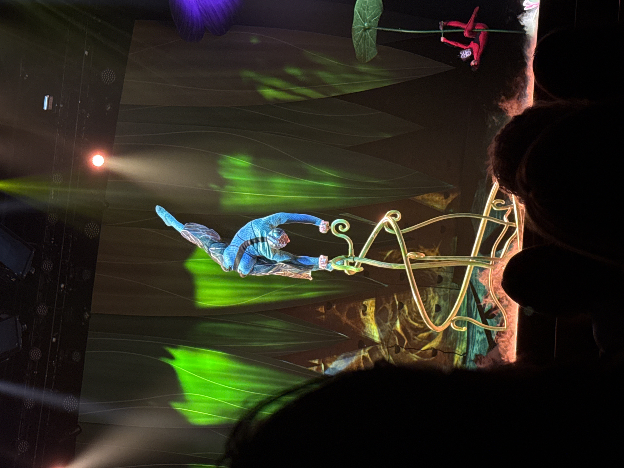 This image depicts an aerial acrobatic performance occurring inside a darkened theater or arena, potentially in Sofia, Bulgaria. The central figure is a performer in a blue and patterned costume, suspended horizontally in mid-air. This individual is actively holding onto a large, ornate, gold-colored prop with swirling, organic designs.
In the upper right portion of the frame, another performer, dressed in a red costume with a stylized face mask, hangs from a green, stem-like rope or bar. The stage background is illuminated with projections of vibrant green leaf patterns and wavy lines, contributing to a fantastical and immersive setting. Dramatic stage lighting, including bright spotlights and LED fixtures, casts beams of white, green, and reddish-orange light across the scene from the left.
The activity showcases the athleticism and artistry of the performers in a choreographed aerial act. Silhouetted heads of audience members are visible in the foreground and along the right edge, indicating a live performance setting. The overall mood is theatrical and visually engaging, characteristic of a circus or aerial show. No text is visible in the image.
