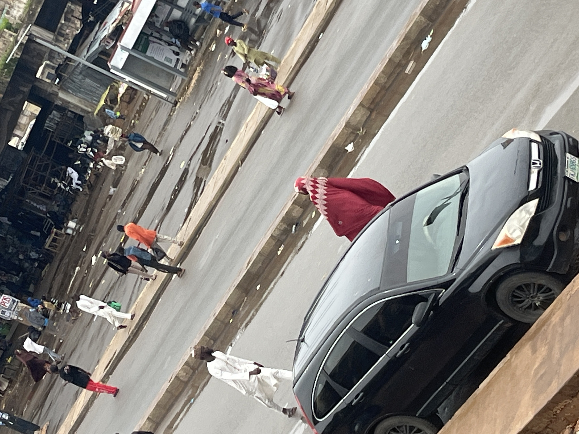In Jos, Nigeria, a black Honda CR-V is parked on a wet asphalt road with its front facing right. Several people are on the road and sidewalk. In the foreground, a person wearing a red hijab and a white and red patterned garment stands near the car. Further down the road, a man in a white traditional outfit walks carrying a green bag. To his right, a man in an orange shirt walks with others. In the background, market stalls and structures line the street. The scene appears to be an outdoor market or busy street.
