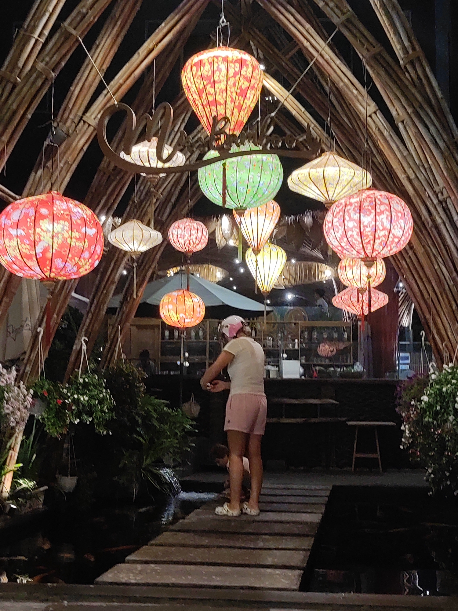 The image depicts an outdoor night scene at a venue, possibly a restaurant or cafe, with a decorative bamboo structure overhead. A variety of colorful, illuminated lanterns, including red, green, and yellow, hang from the structure, creating a festive and warm ambiance. In the foreground, a person wearing a white t-shirt, pink shorts, and a pink helmet stands on a wooden walkway, facing away from the camera. The walkway appears to lead over a dark body of water, possibly a pond or water feature, with lush greenery and some flowering plants on the sides. In the background, shelves with bottles are visible, suggesting a bar or service area, and a light blue umbrella provides shade. The scene is illuminated by the lanterns and ambient light, indicating it is nighttime. The overall mood is relaxed and inviting. The word "ONE" is visible in cursive script on a metal sign hanging from the bamboo structure.