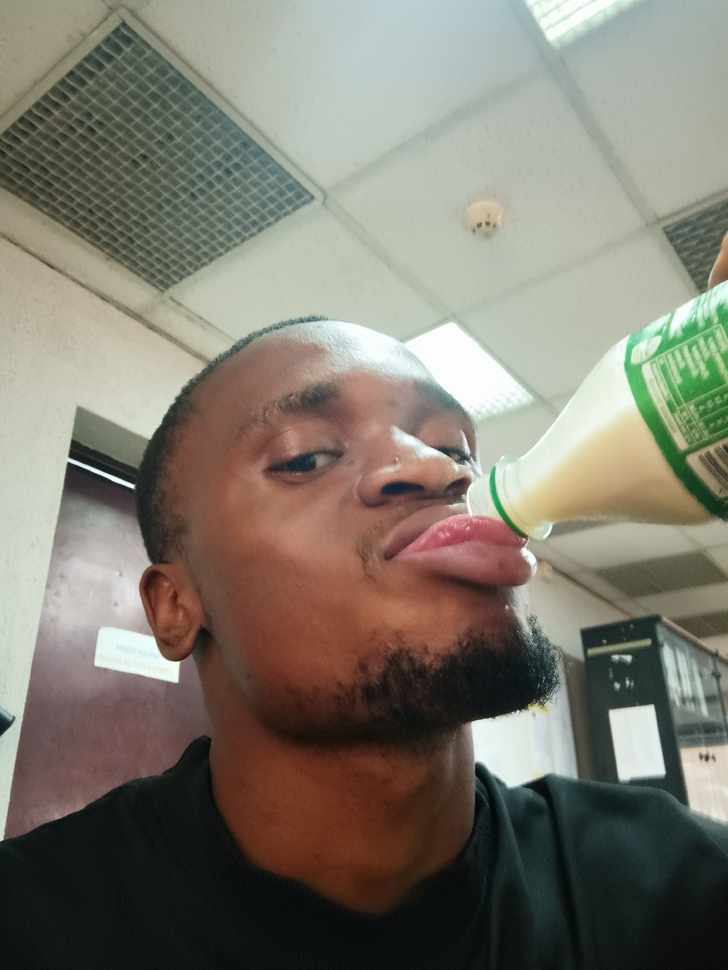 An eye-level indoor shot captures a Black man with short dark hair and a beard, wearing a black collared shirt, looking directly at the camera while drinking. His lips are puckered around the opening of a plastic bottle, and his tongue is slightly extended, indicating he is actively consuming a light-colored liquid, possibly a dairy beverage. The bottle has a green label with small, indistinct white text and a partially visible barcode.
The setting is an indoor space, likely an office or institutional building in Kakuri Gwari, Nigeria, suggested by the overhead infrastructure. The ceiling features white grid tiles, with a square ventilation grille visible on the left and a round smoke detector and a fluorescent light fixture towards the center-right. To the man's left, in the background, is a dark door or wall with a white sign attached. The text on this sign is blurry, but "MOOR EVER", "ROOM", and "ONE" might be discernible. To the far right, a dark, tall piece of equipment or furniture is partially visible. The lighting is artificial, and the man's expression appears casual and focused on his drink.