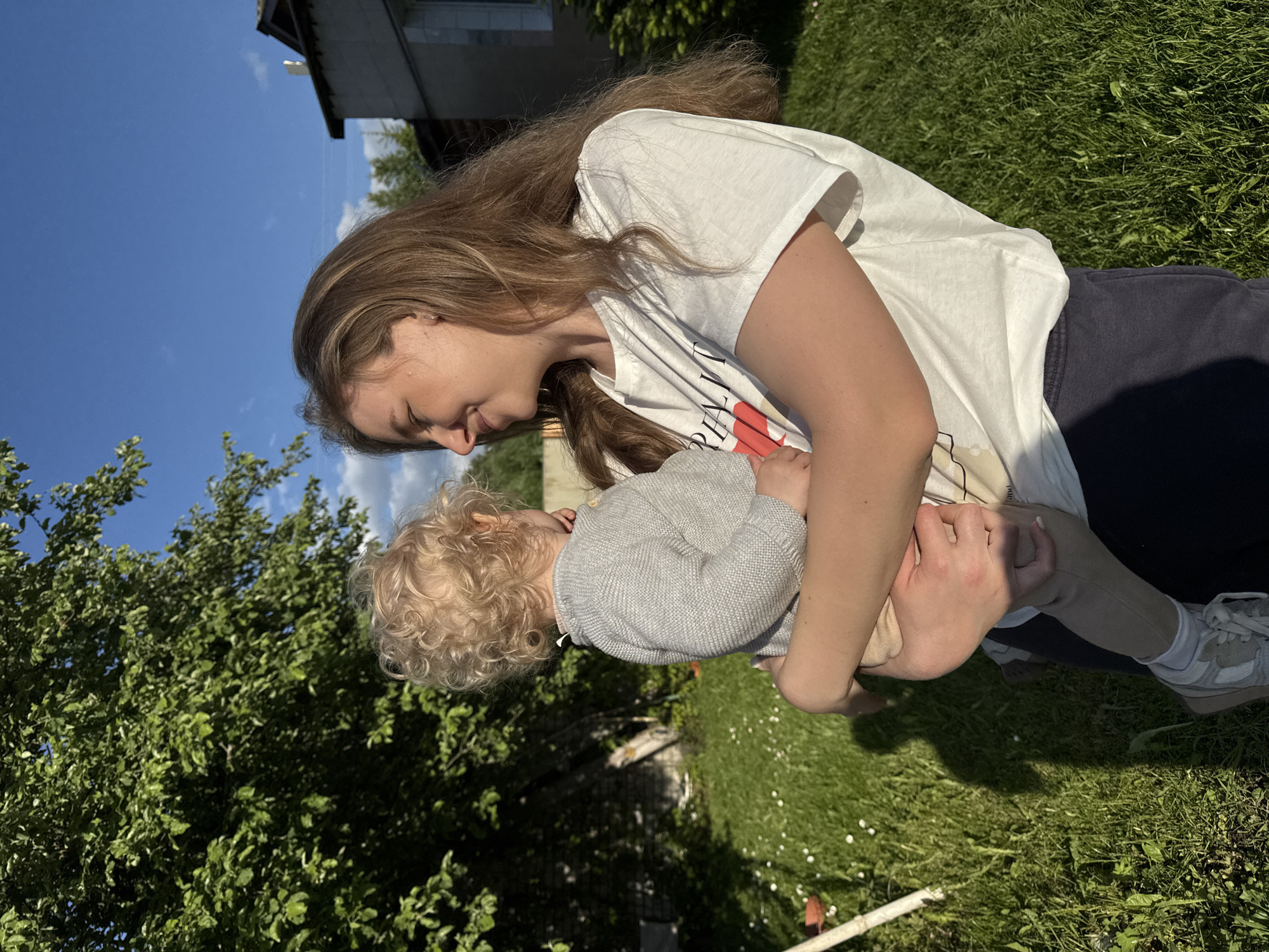 The image, taken in Красный Маяк, Russia, depicts a tender moment between a young woman and a small child outdoors on a sunny day. The scene is set in a vibrant green, grassy yard, with lush leafy trees providing a natural backdrop and a building visible in the upper background against a clear blue sky with scattered white clouds.
The young woman, with long, light brown hair, is seen from the side, gently looking down at the child she holds. She wears a white t-shirt with a graphic design that includes partially visible text such as "THE ONLY" and "TREND", and dark blue pants. She has light-colored sneakers paired with white socks. Her expression conveys warmth and affection towards the child.
The small child, with distinctive curly blonde hair, is dressed in a light grey knitted cardigan, which features small yellow accents near the shoulder, and light-colored pants. The child is looking up towards the woman, suggesting an engaged and loving interaction.
The strong sunlight creates noticeable shadows on the grass, indicating a bright and clear day. The overall impression is one of a peaceful, caring moment shared between an adult and a child in a natural, outdoor setting.