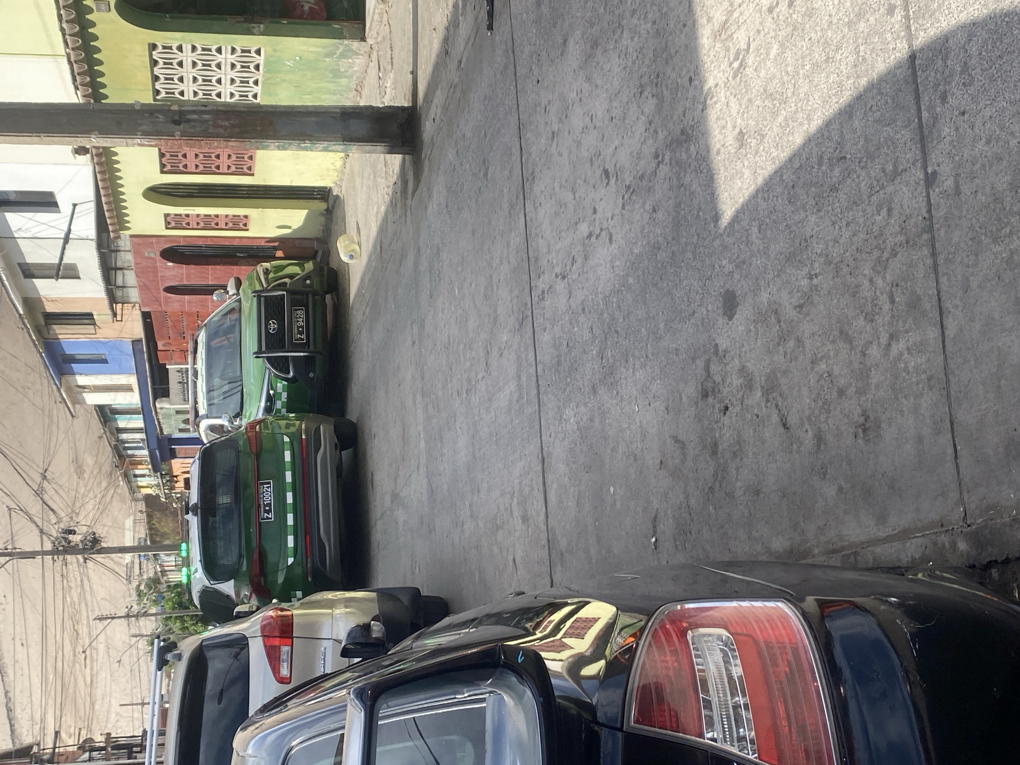 The image shows a street scene in Arica, Chile. The view is looking down a narrow street with buildings on the left and a sidewalk on the right. Several cars are parked along the left side of the street. A green car with license plate Z*9428 and a similar green car with plate Z*10021 can be seen among the parked vehicles. Power lines run along the left side of the street. Buildings are visible in the background. The street surface is concrete. The lighting suggests it is daytime. The black car is in the foreground.